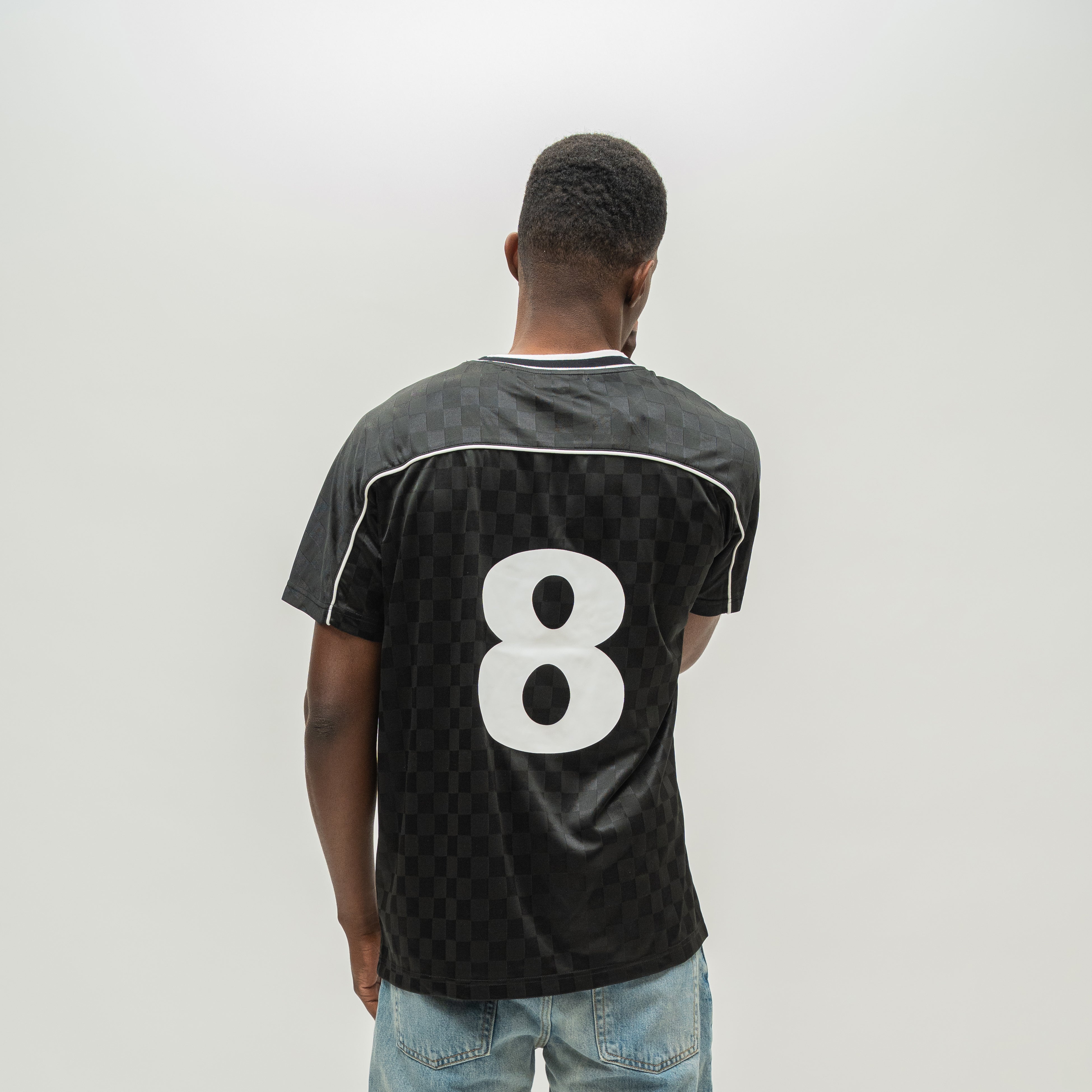 A man viewed from behind wearing a black checkered soccer jersey with the number 8 on the back, paired with light blue jeans, against a plain light gray background.