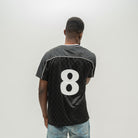 A man viewed from behind wearing a black checkered soccer jersey with the number 8 on the back, paired with light blue jeans, against a plain light gray background.