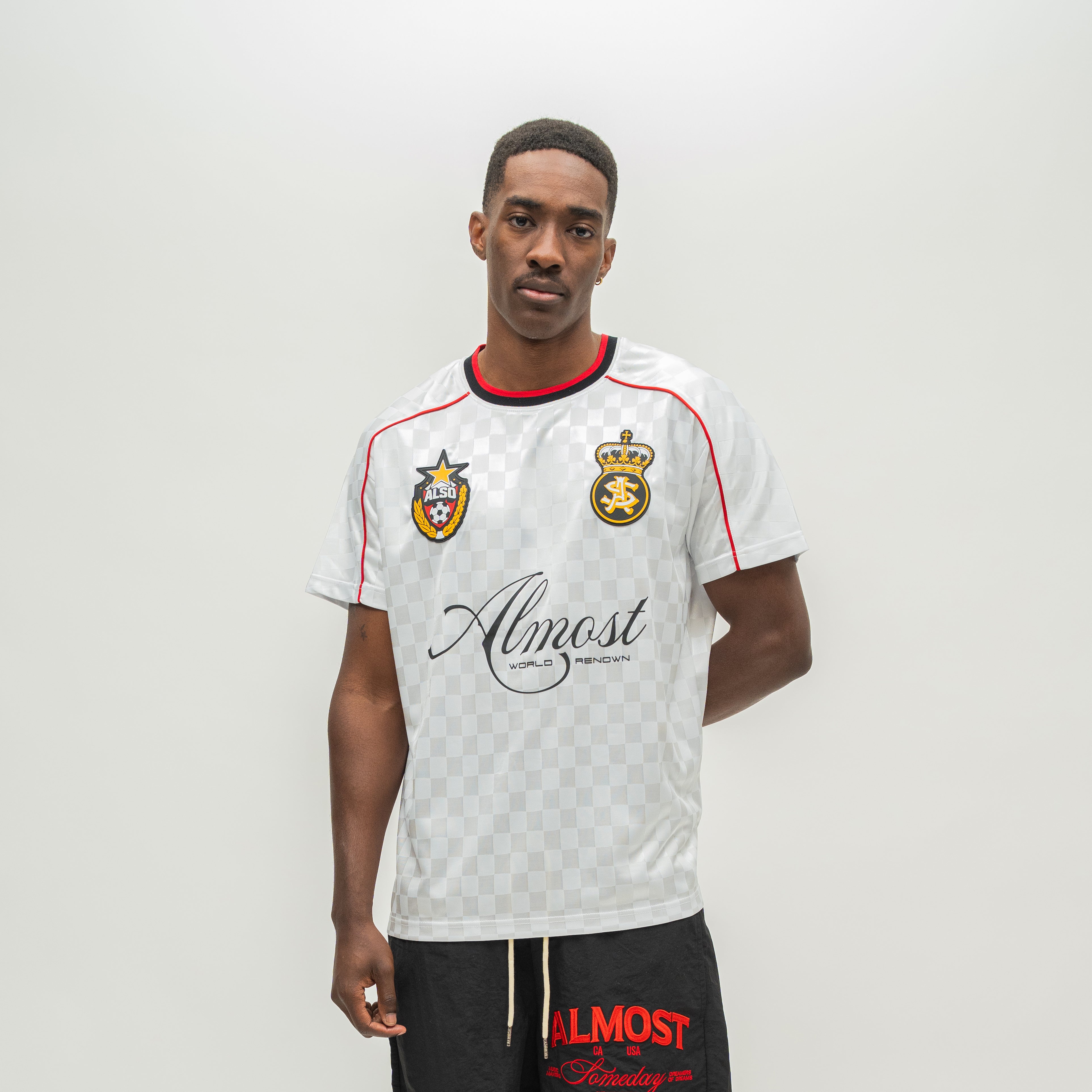 A man modeling a white checkered short-sleeve soccer jersey with red and black trim, featuring a crest and 'Almost Someday' sponsor logo, paired with black shorts.