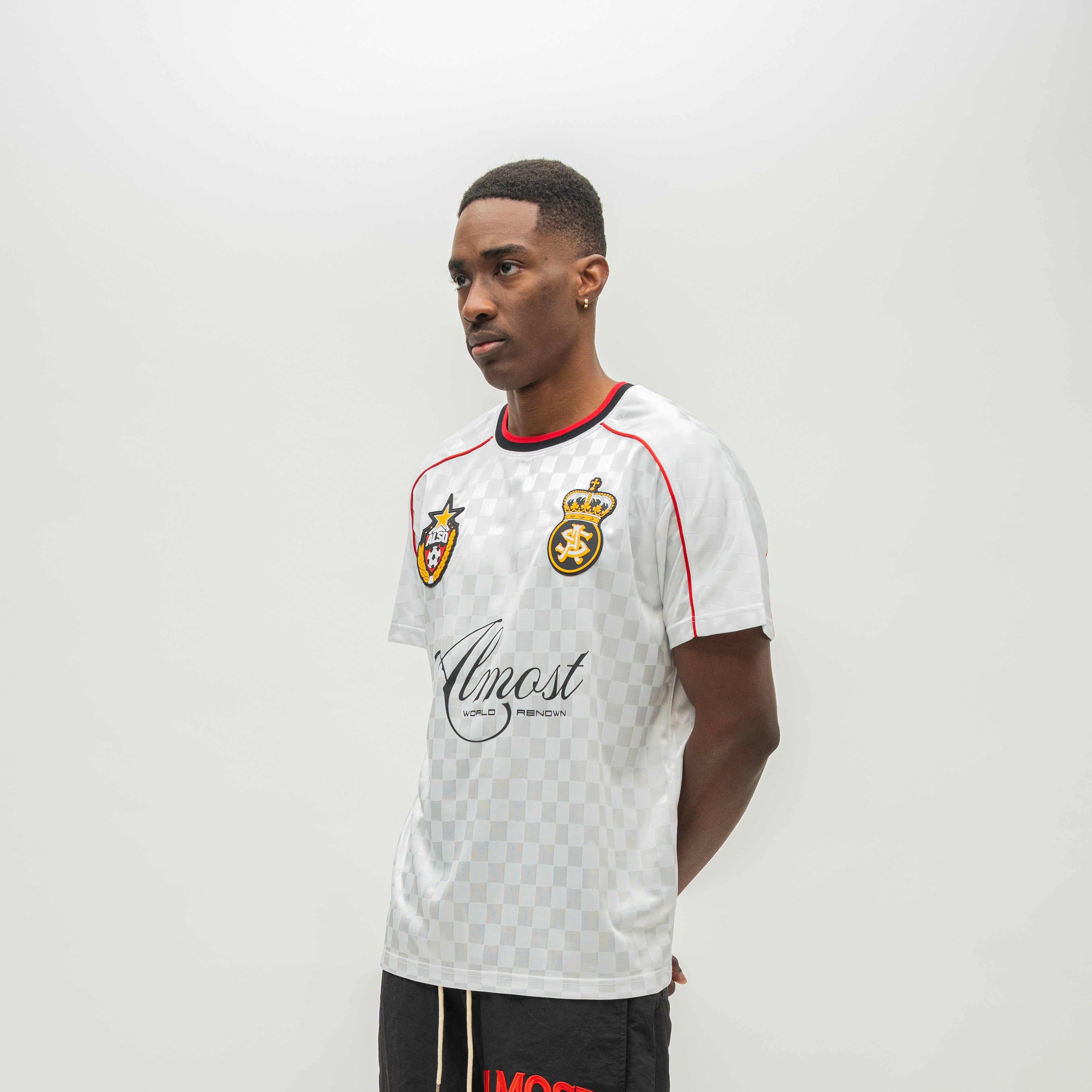 A man modeling a white checkered soccer jersey with red and navy trim, featuring the Almost Someday brand logo on the front, set against a plain light gray background.