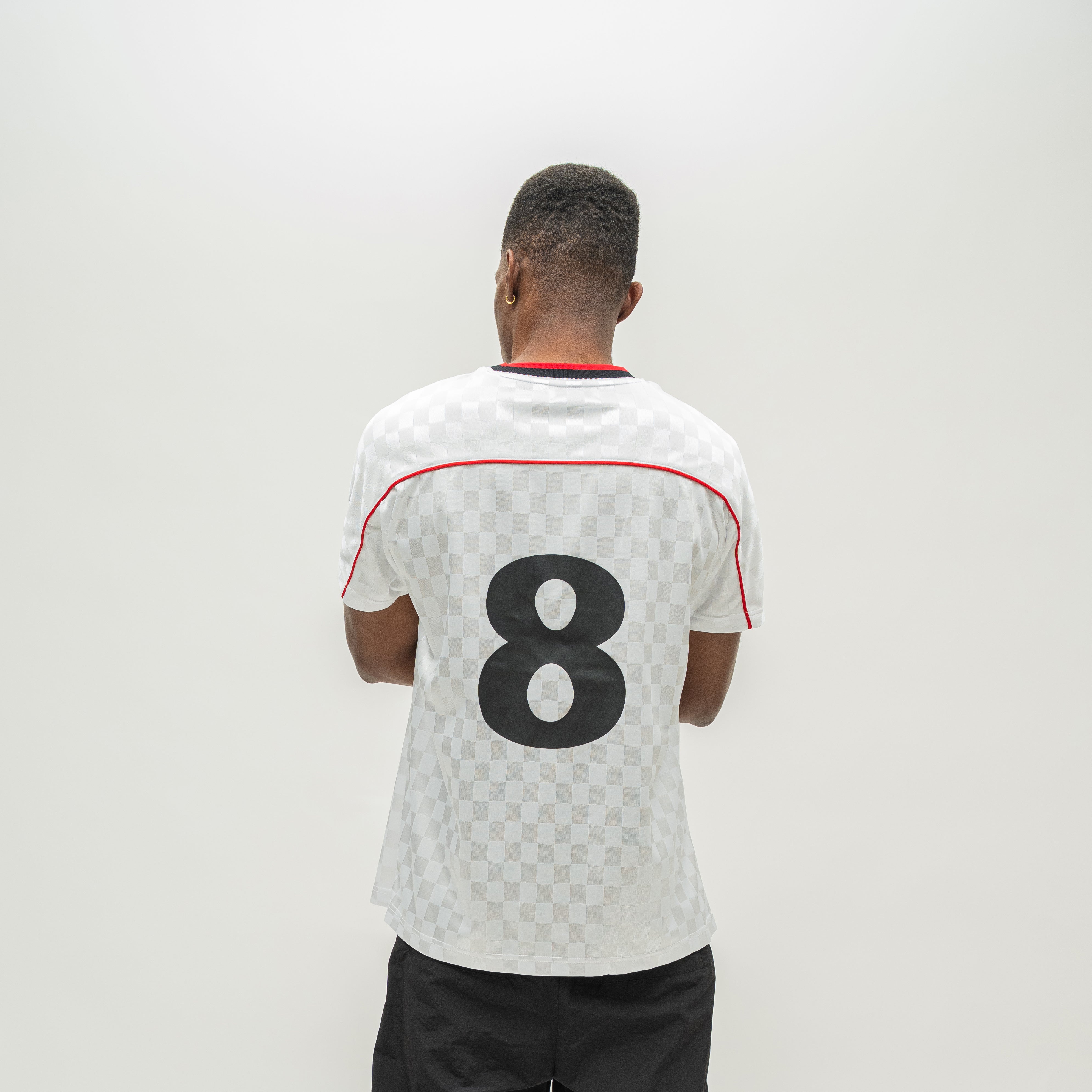 A man viewed from behind wearing a white soccer jersey with a black and white checkerboard pattern, a red collar trim, and the number 8 printed in black on the back.