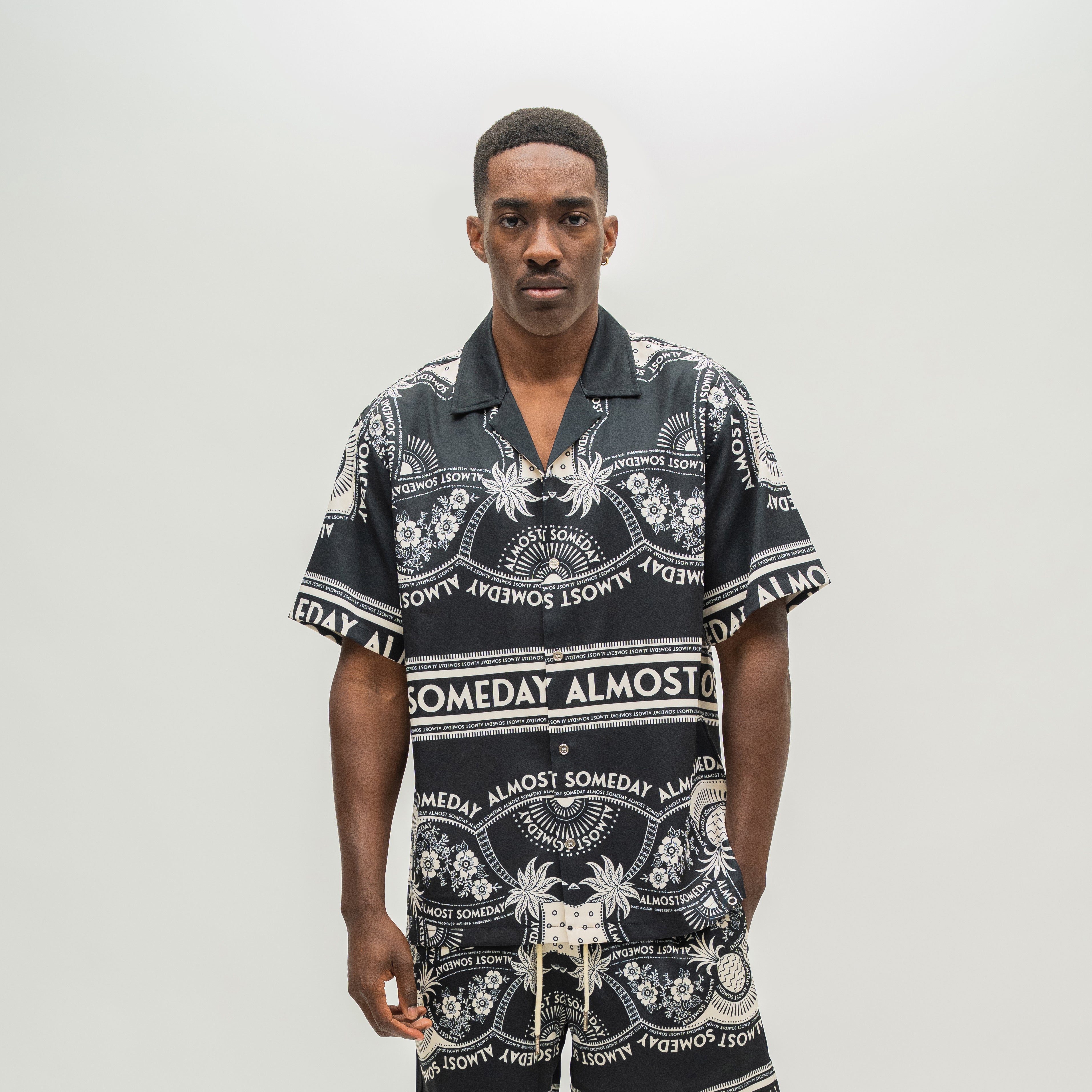A man modeling a matching black and white patterned short-sleeve button-up shirt and shorts set. The shirt features a bold graphic print with the words 'ALMOST SOMEDAY' and tropical motifs.