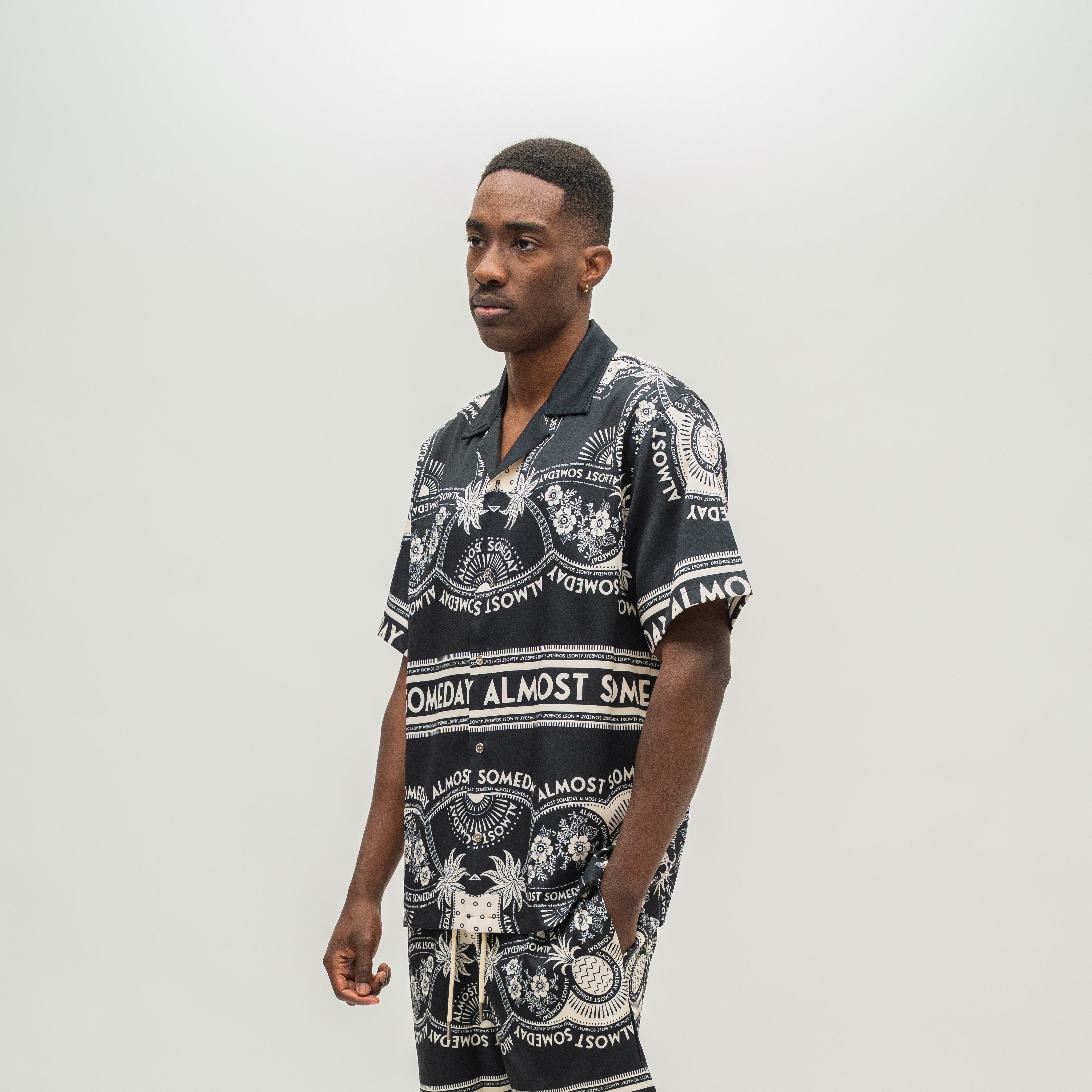 A man wearing a black short-sleeve button-up shirt with a white graphic print, standing against a light gray studio background.