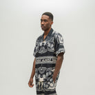 A man wearing a black short-sleeve button-up shirt with a white graphic print, standing against a light gray studio background.
