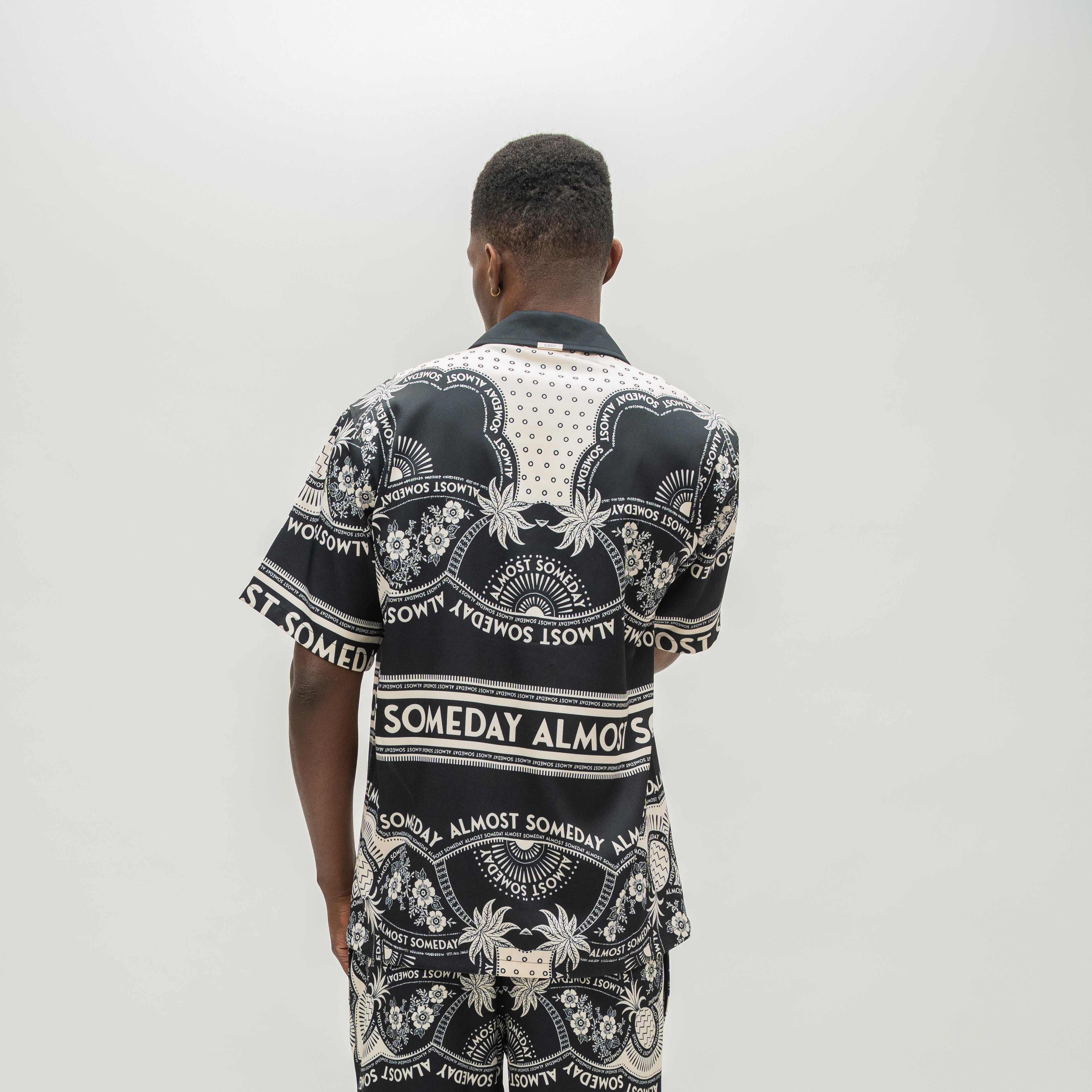 A man viewed from behind wearing a black and white patterned short-sleeve button-up shirt with 'Almost Someday' text and ornate graphic motifs, against a plain light gray background.