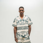 A man wearing a matching short-sleeve camp collar shirt and shorts set with a cream base and dark green graphic print featuring palm trees and the text 'ALMOST SOMEDAY'.