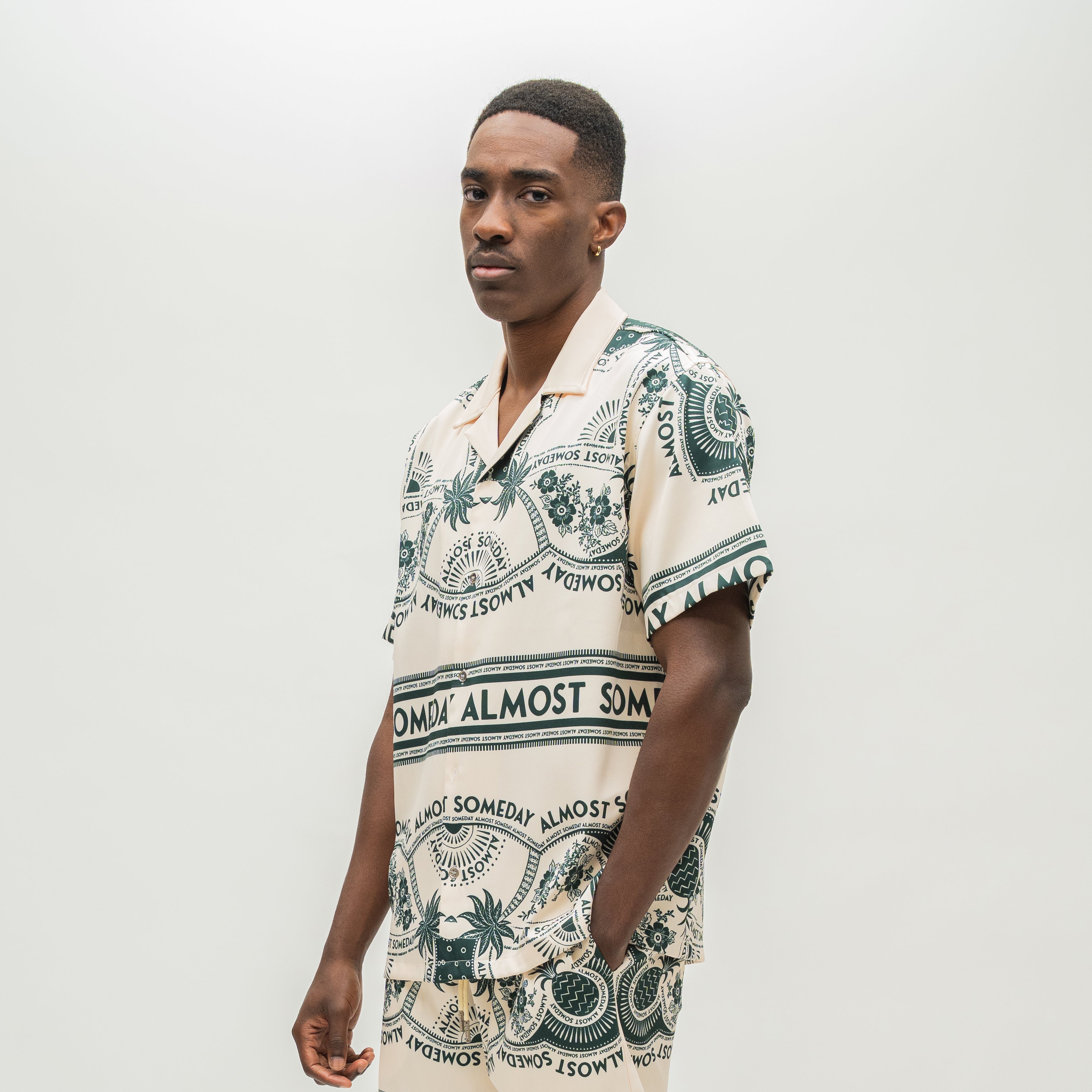 A man wearing a short-sleeve camp collar shirt with a cream base and dark green tropical-themed print featuring palm trees, pineapples, and the text 'ALMOST SOMEDAY'.