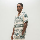 A man wearing a short-sleeve camp collar shirt with a cream base and dark green tropical-themed print featuring palm trees, pineapples, and the text 'ALMOST SOMEDAY'.