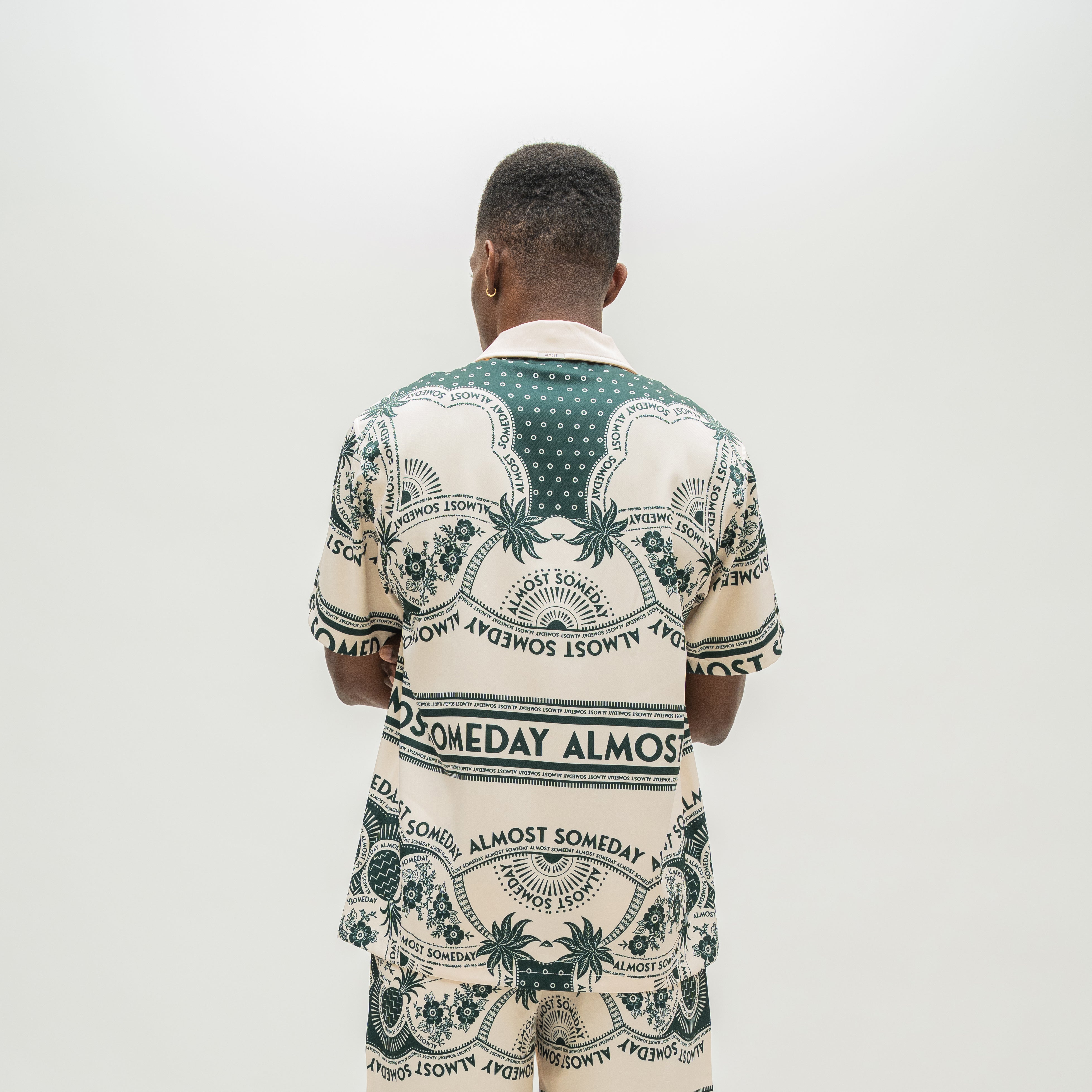 A man viewed from behind wearing a matching short-sleeve button-up shirt and shorts set in cream with a dark green graphic print featuring palm trees and the text 'ALMOST SOMEDAY'.