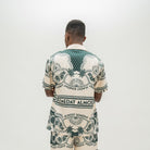 A man viewed from behind wearing a matching short-sleeve button-up shirt and shorts set in cream with a dark green graphic print featuring palm trees and the text 'ALMOST SOMEDAY'.