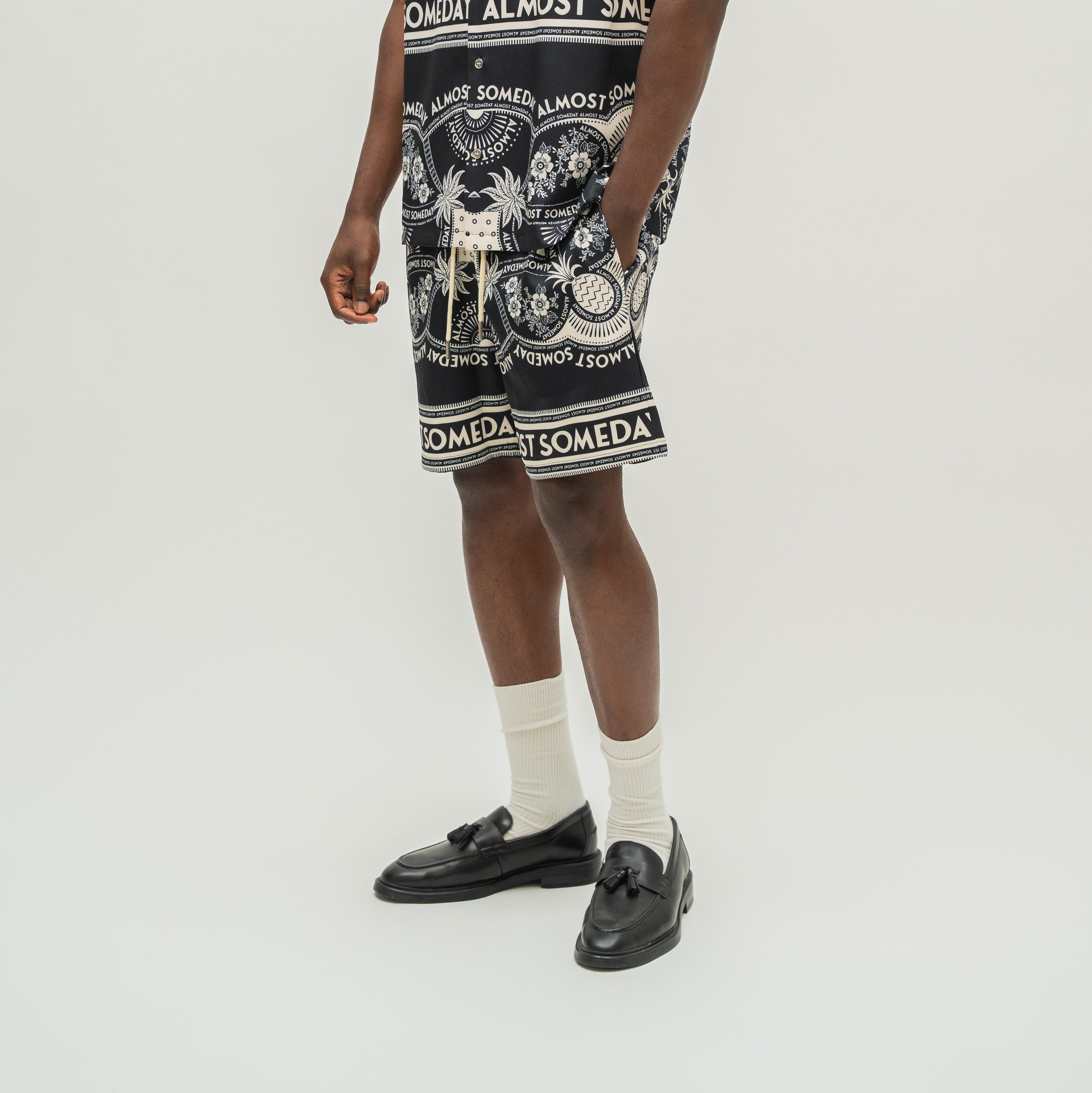 A model wearing black and white patterned shorts with 'ALMOST SOMEDAY' branding, styled with white socks and black leather loafers against a light gray studio background.