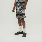 A model wearing black and white patterned shorts with 'ALMOST SOMEDAY' branding, styled with white socks and black leather loafers against a light gray studio background.