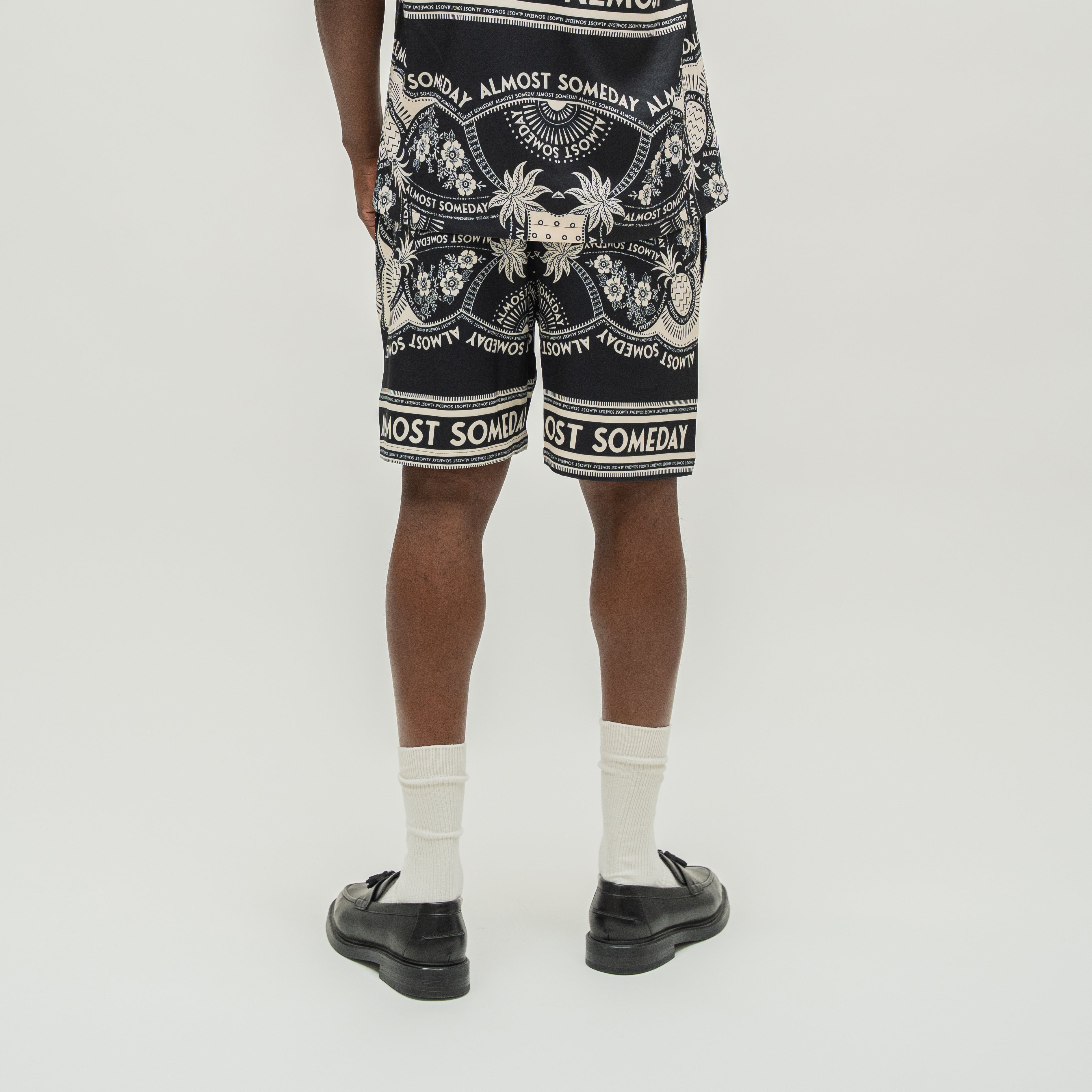 A person shown from the back wearing black shorts with a white ornate graphic print and text, paired with white socks and black loafers, against a light gray studio background.