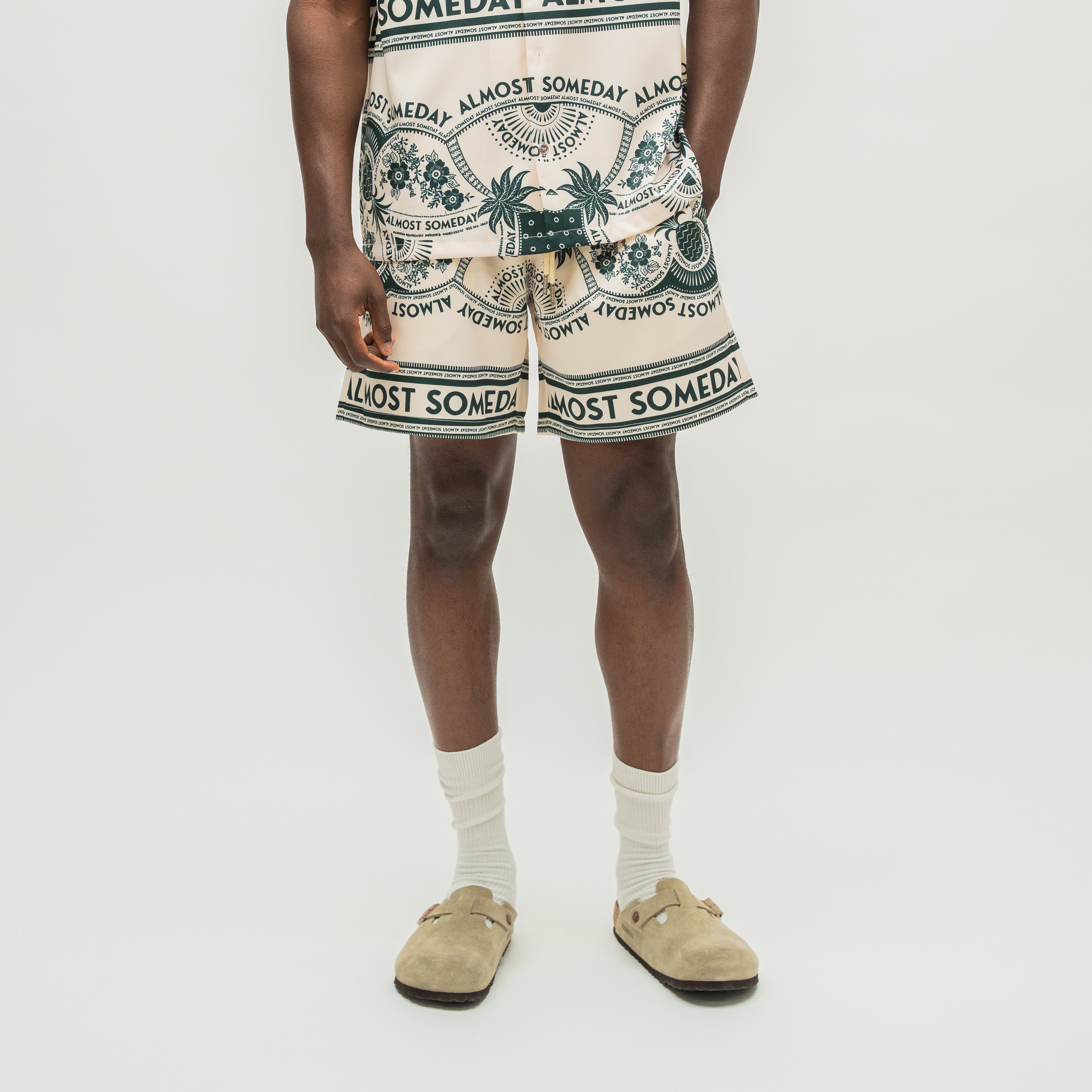 A model is shown from the waist down wearing cream-colored shorts with a dark green and black tropical print and text, paired with white socks and beige suede clogs, against a plain light gray background.