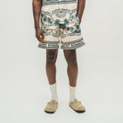 A model is shown from the waist down wearing cream-colored shorts with a dark green and black tropical print and text, paired with white socks and beige suede clogs, against a plain light gray background.