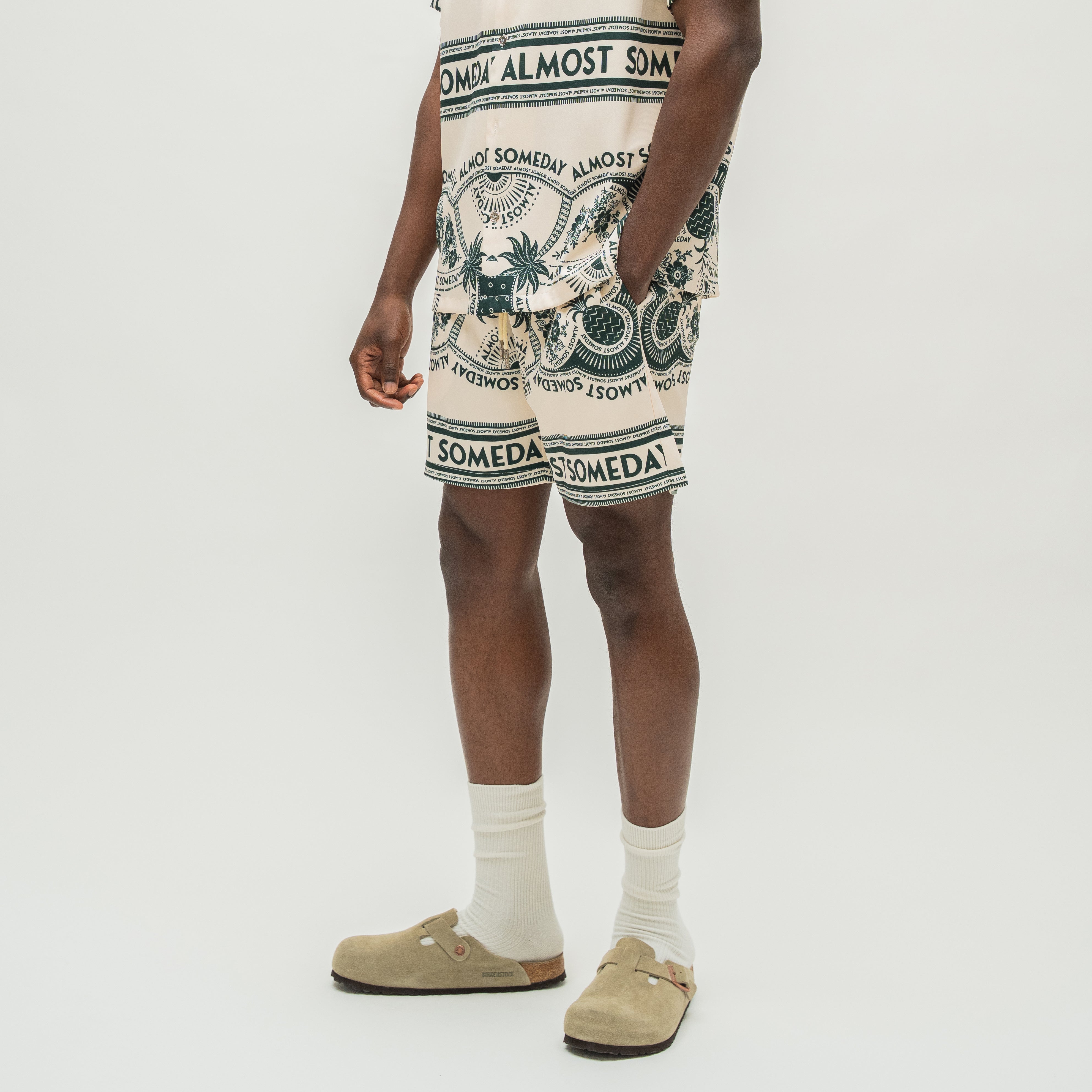 A model wearing a beige and navy blue patterned Almost Someday shorts set, including a matching top and shorts, styled with white socks and beige Birkenstock clogs against a plain white studio background.