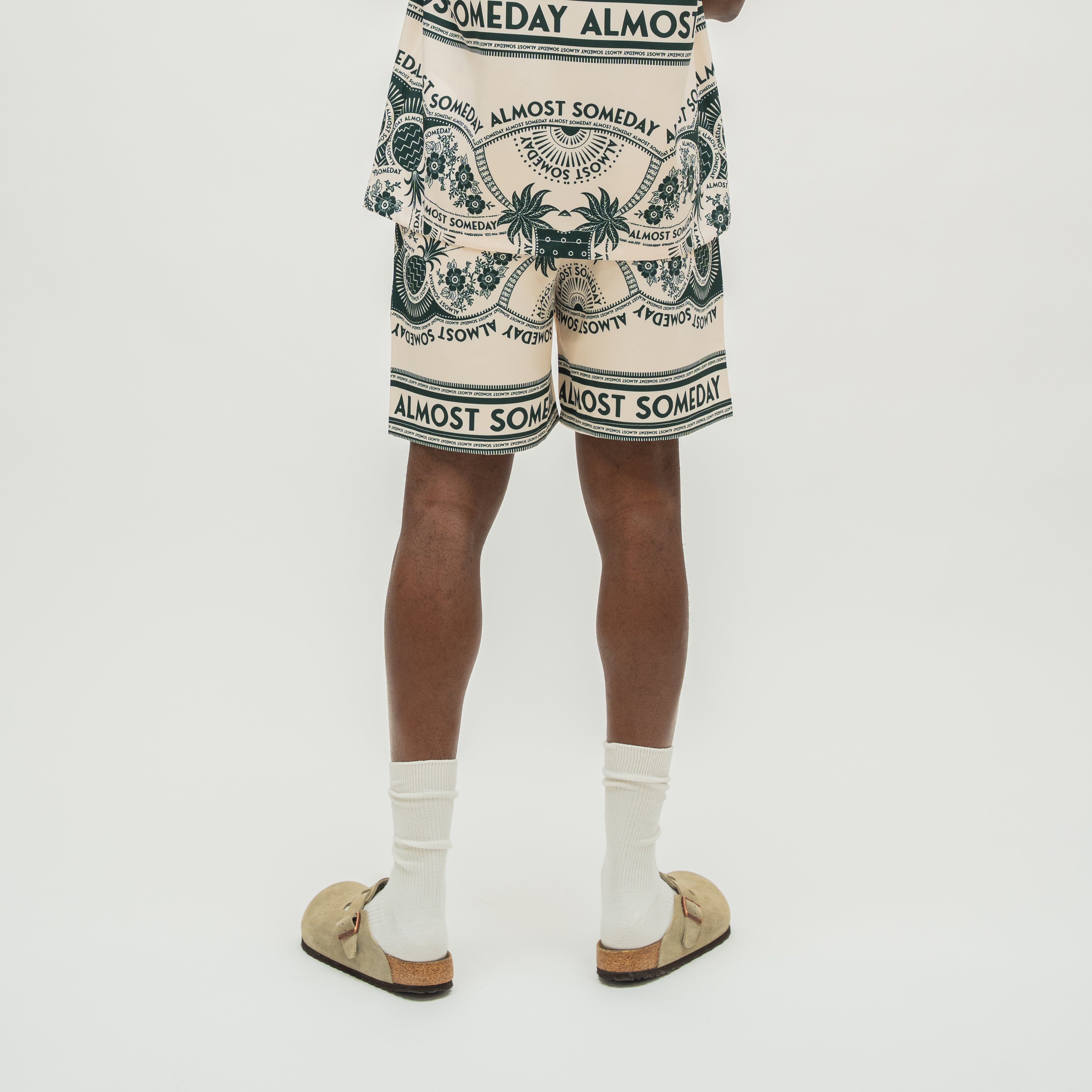 A person shown from the waist down wearing cream-colored shorts with a dark green graphic print and the words 'ALMOST SOMEDAY' repeated around the border. The person is also wearing white socks and tan slide sandals, standing against a plain light gray studio background.