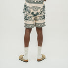 A person shown from the waist down wearing cream-colored shorts with a dark green graphic print and the words 'ALMOST SOMEDAY' repeated around the border. The person is also wearing white socks and tan slide sandals, standing against a plain light gray studio background.
