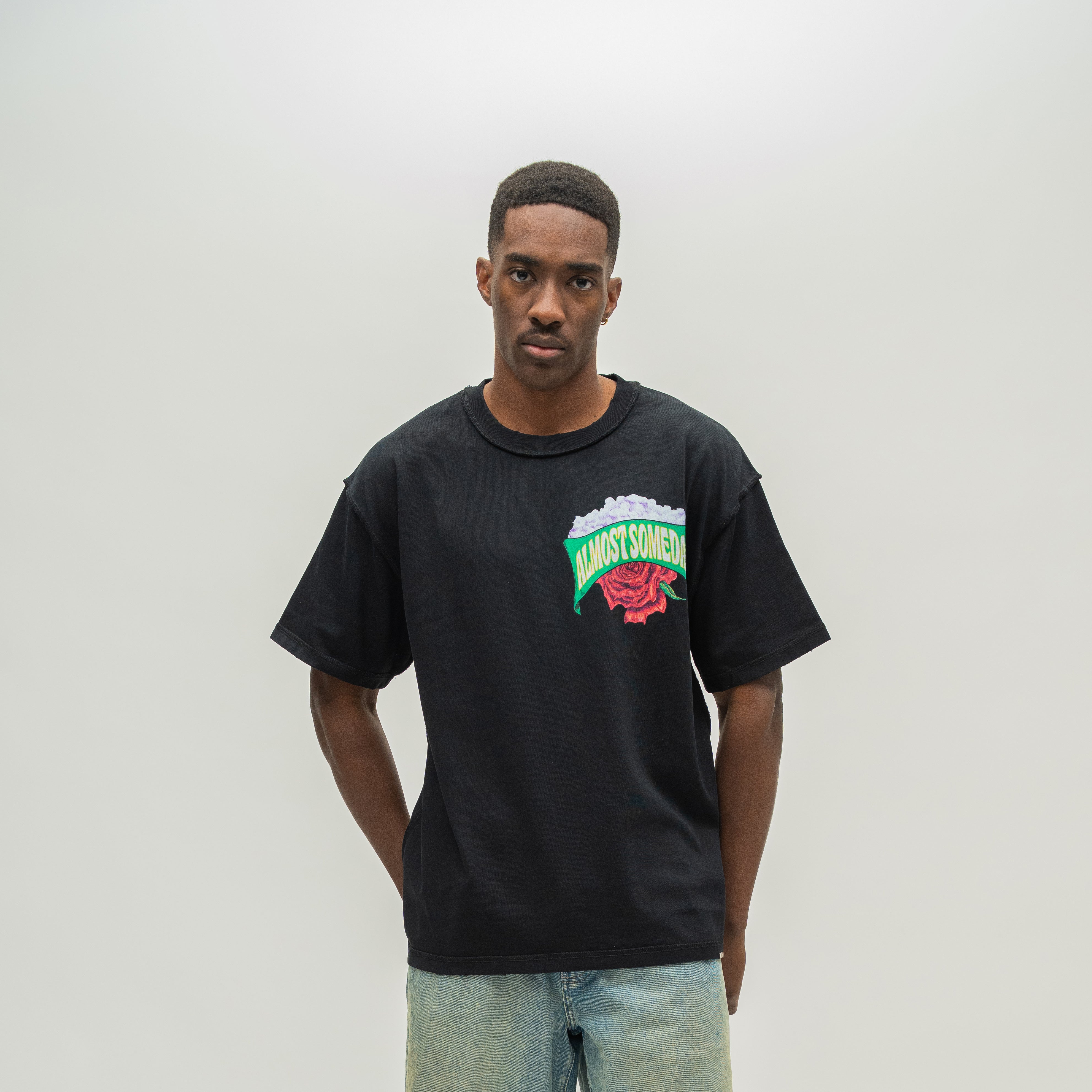 A man wearing a black oversized t-shirt with a colorful 'Almost Someday' graphic on the chest, paired with light wash jeans, standing against a neutral light gray background.