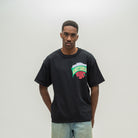 A man wearing a black oversized t-shirt with a colorful 'Almost Someday' graphic on the chest, paired with light wash jeans, standing against a neutral light gray background.