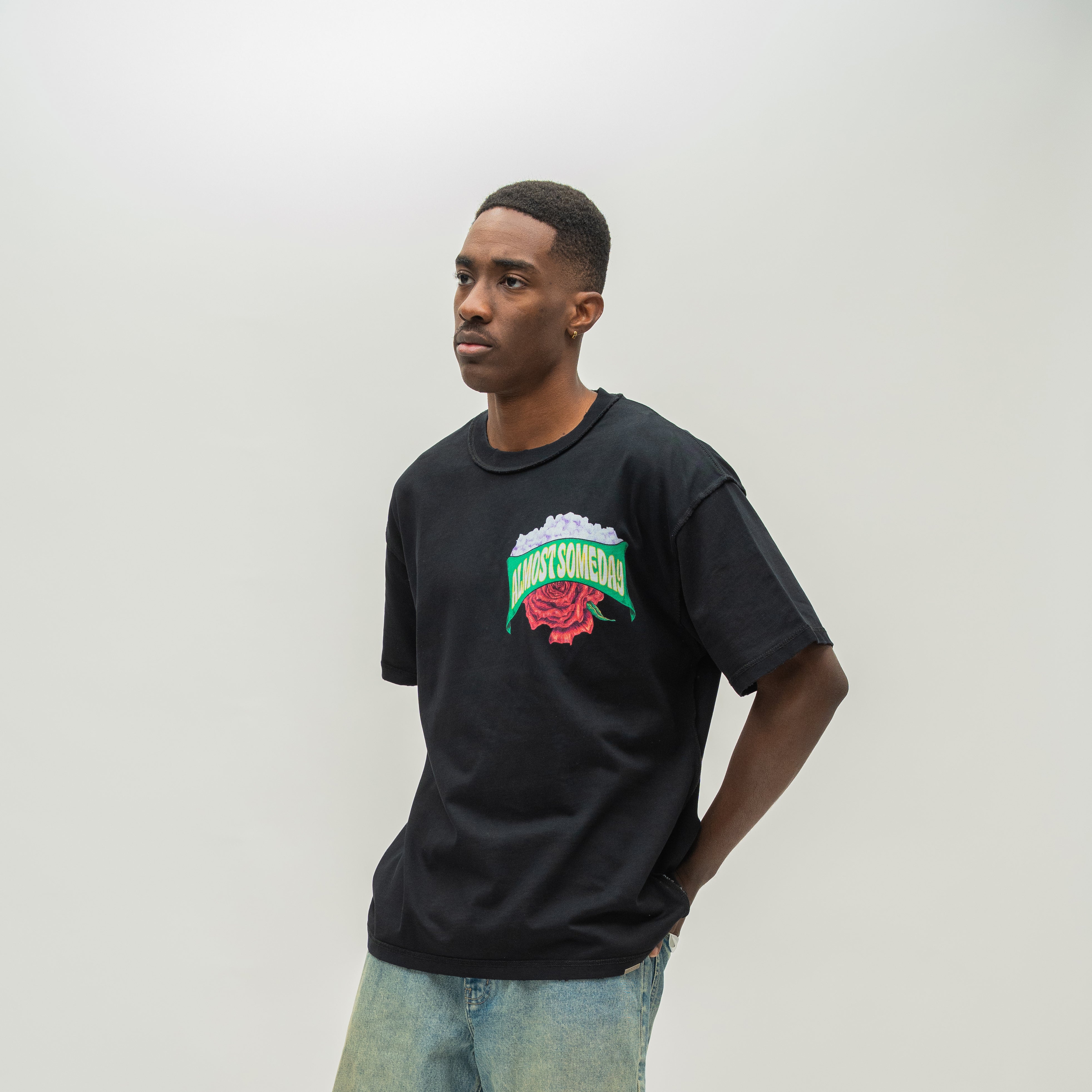 A man wearing a black t-shirt with a colorful 'Almost Someday' graphic on the chest, paired with light blue jeans, standing against a plain light gray background.