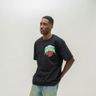 A man wearing a black t-shirt with a colorful 'Almost Someday' graphic on the chest, paired with light blue jeans, standing against a plain light gray background.
