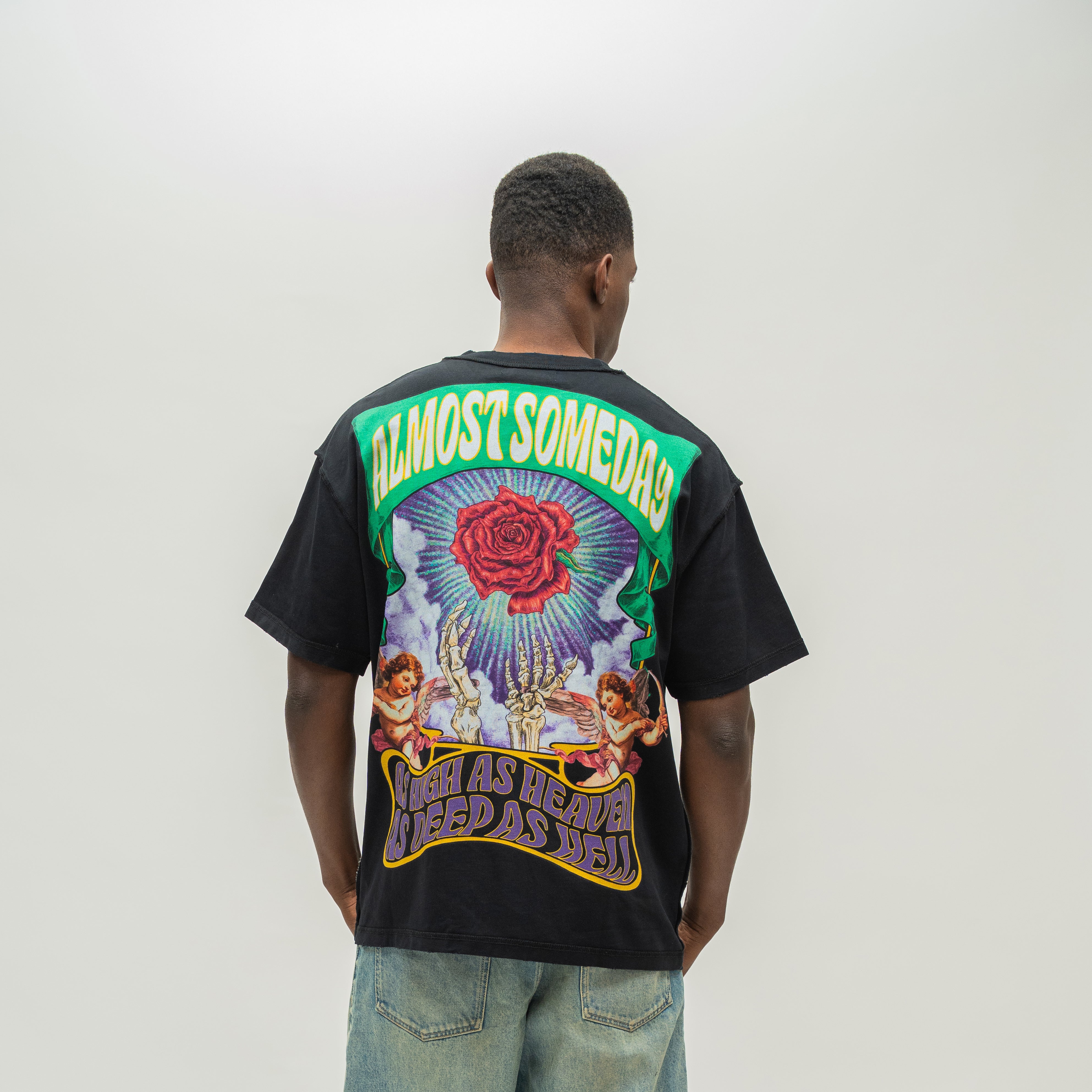 A man viewed from behind wearing a black graphic t-shirt with a large colorful print on the back, paired with light blue jeans, against a plain light gray studio background.