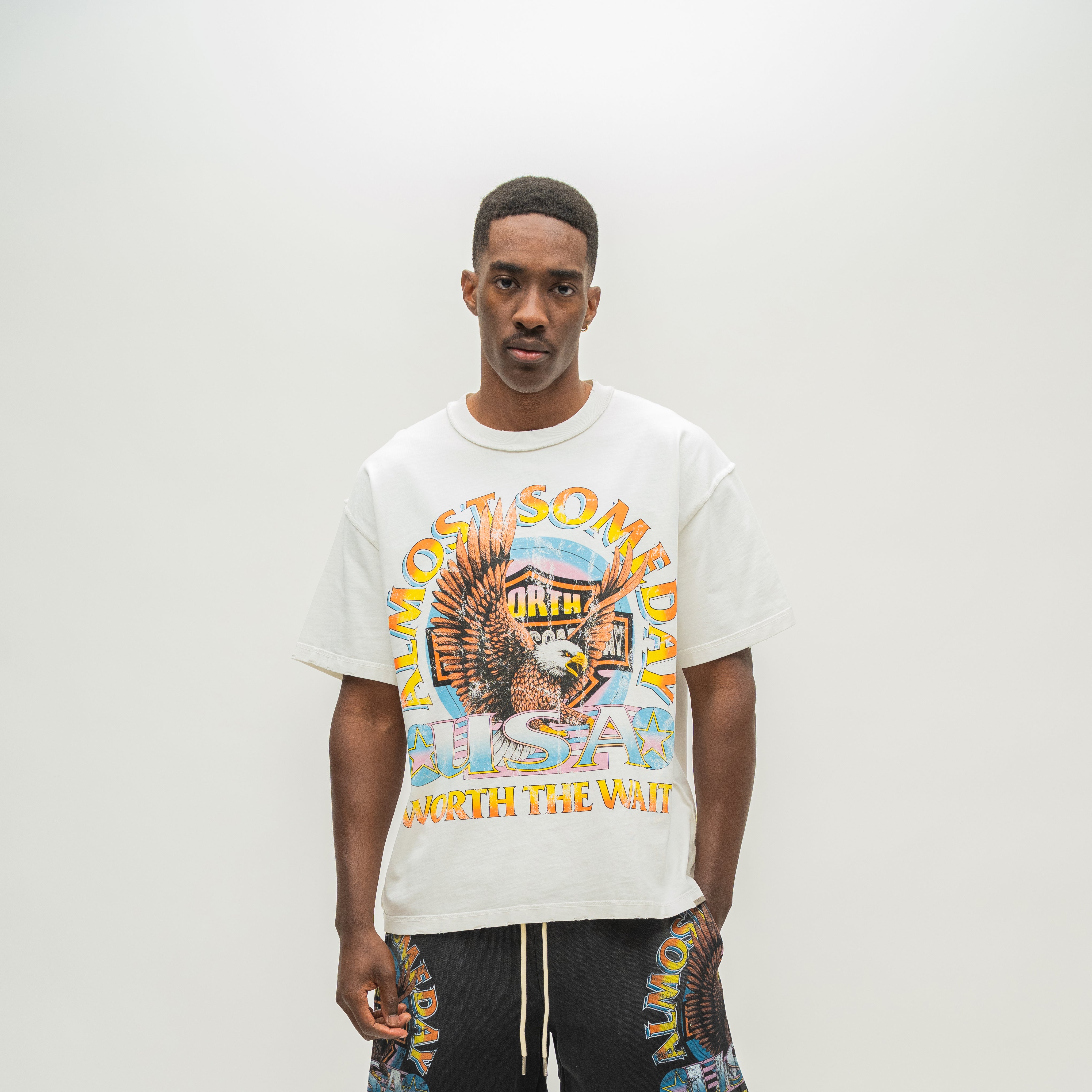 A man wearing a white graphic t-shirt with a colorful 'Almost Someday' and 'USA Worth the Wait' design, paired with black drawstring shorts, against a plain light gray background.