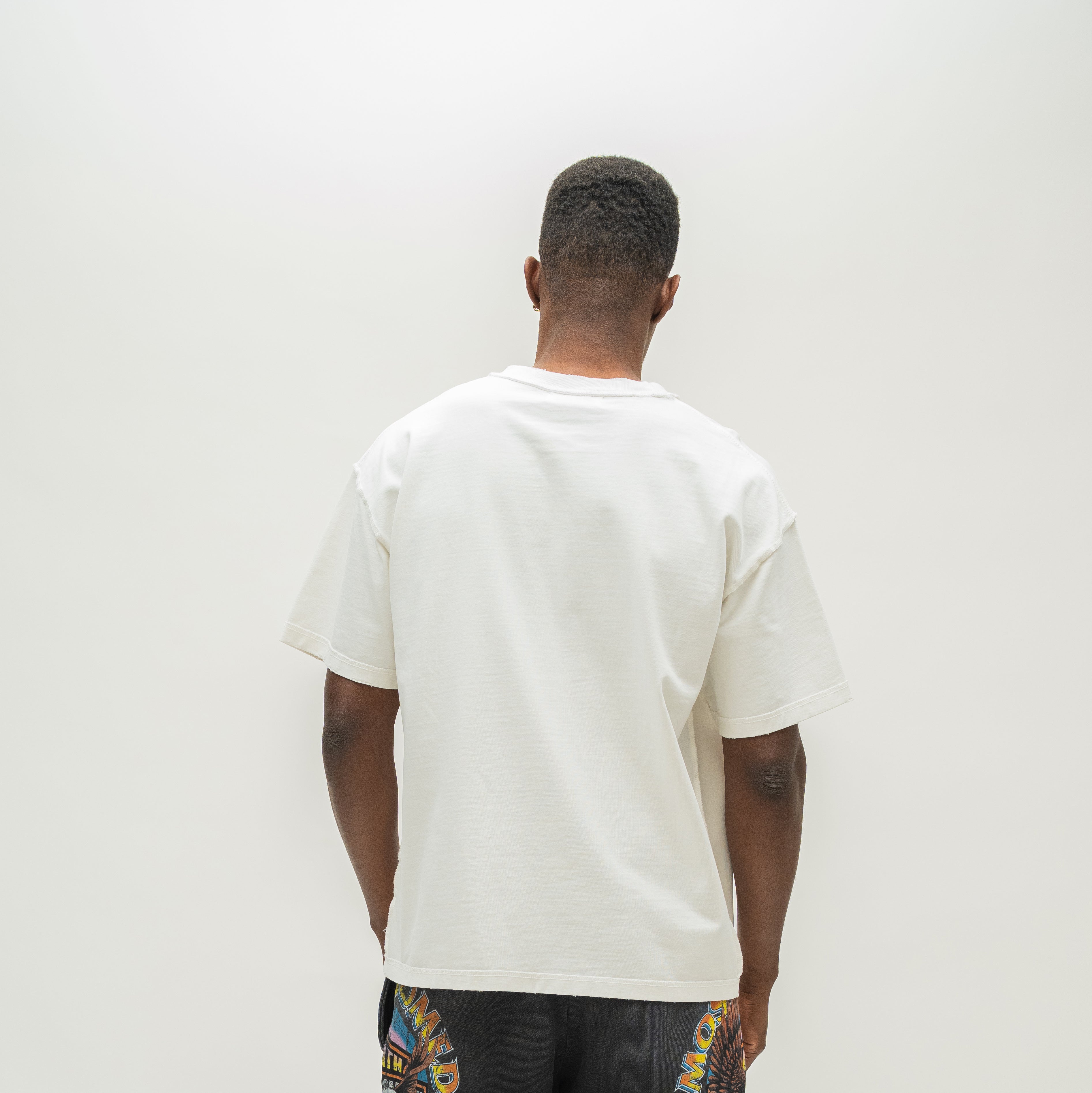 A man viewed from behind wearing an off-white short-sleeve t-shirt and dark patterned shorts against a plain white background.