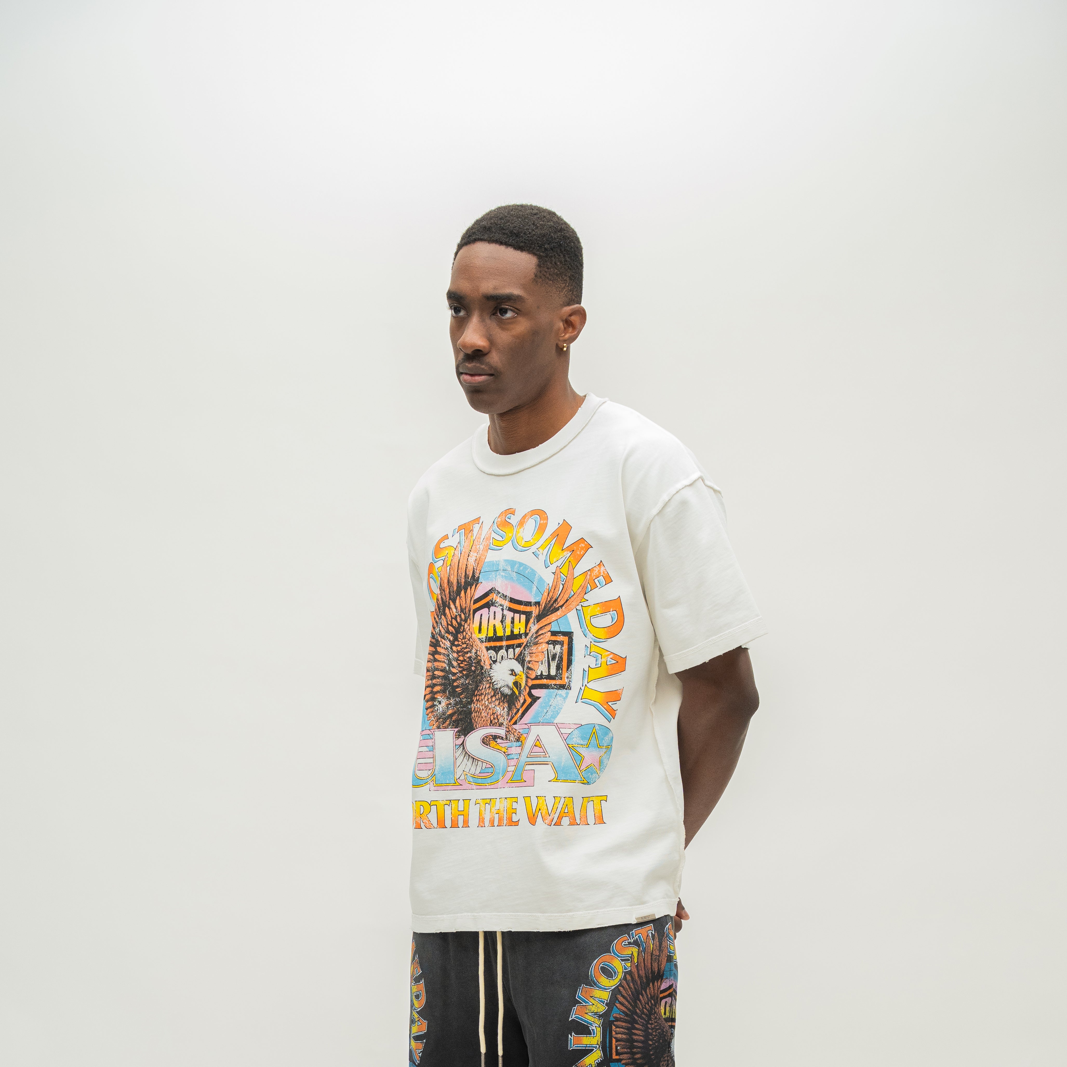 A man wearing a cream-colored graphic t-shirt with a colorful 'Almost Someday USA' design, paired with dark gray shorts, standing against a plain off-white studio background.