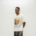 A man wearing a cream-colored graphic t-shirt with a colorful 'Almost Someday USA' design, paired with dark gray shorts, standing against a plain off-white studio background.