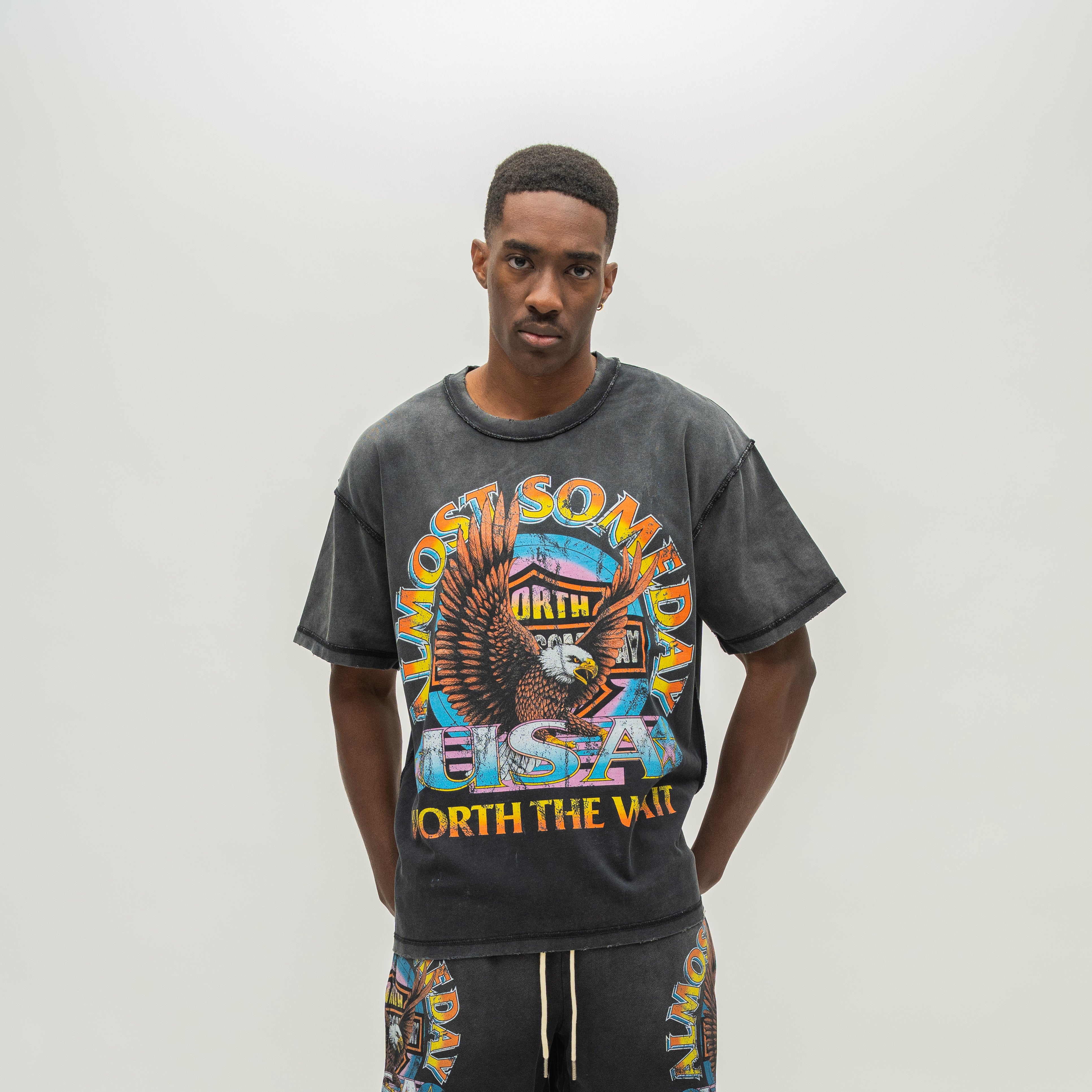 A man wearing a dark gray acid-wash graphic t-shirt with a colorful front print that reads 'ALMOST SOMEDAY' and 'WORTH THE WAIT', paired with matching dark gray sweatshorts, against a plain light gray background.