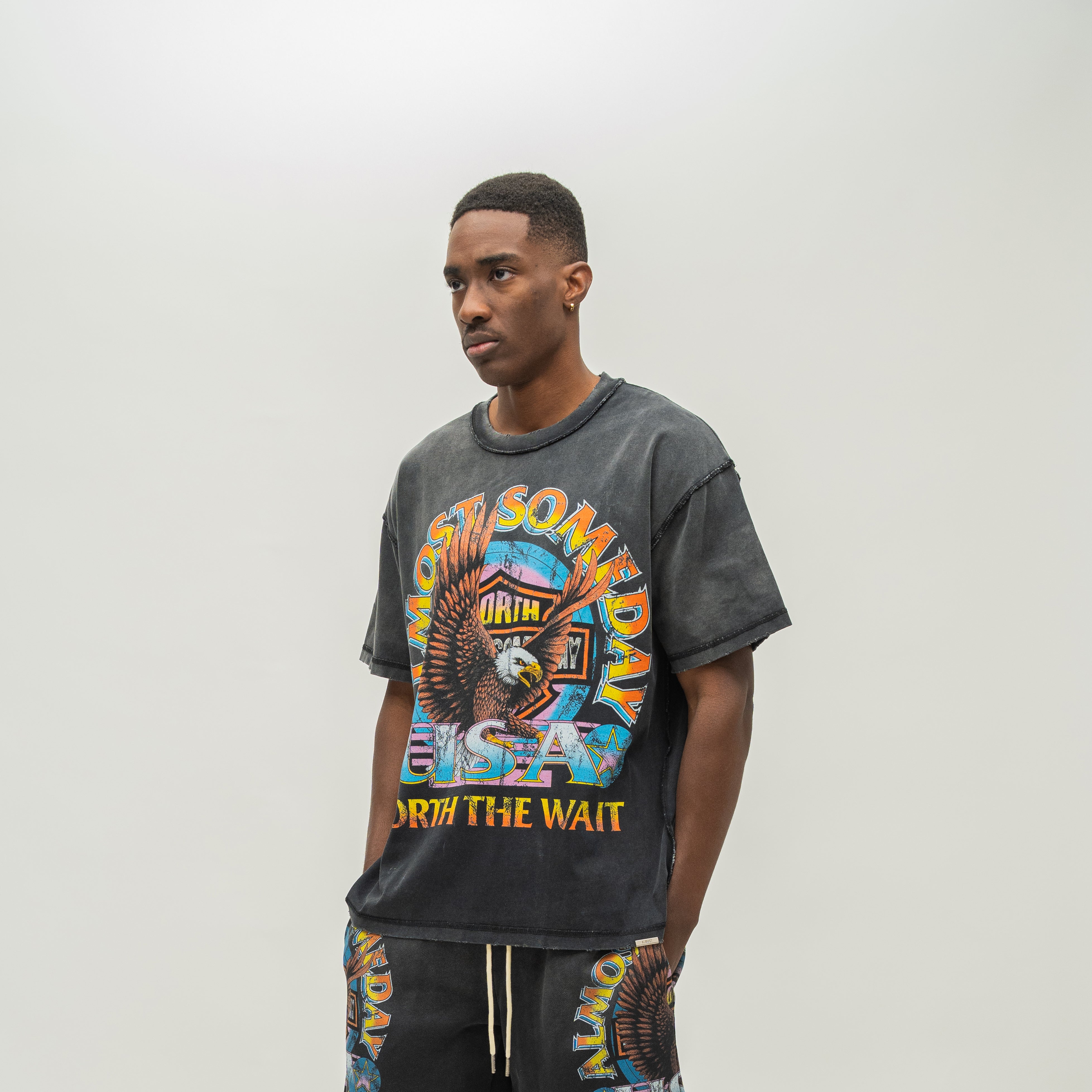 A man wearing a washed black graphic t-shirt and matching sweatpants with a colorful 'Almost Someday' design, standing against a plain light gray studio background.