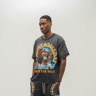 A man wearing a washed black graphic t-shirt and matching sweatpants with a colorful 'Almost Someday' design, standing against a plain light gray studio background.