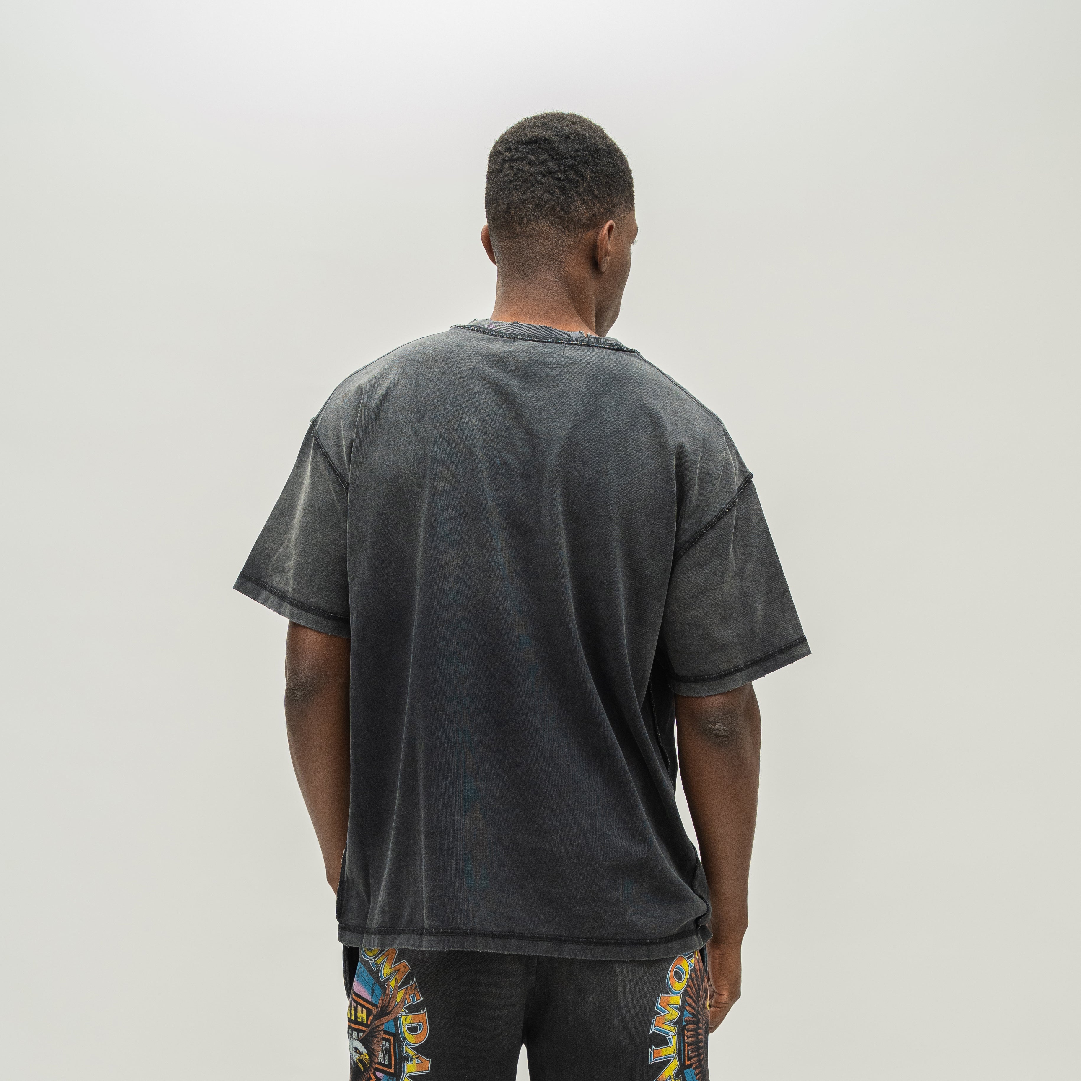 A man viewed from behind wearing an oversized faded black t-shirt and patterned black shorts against a plain light gray background.
