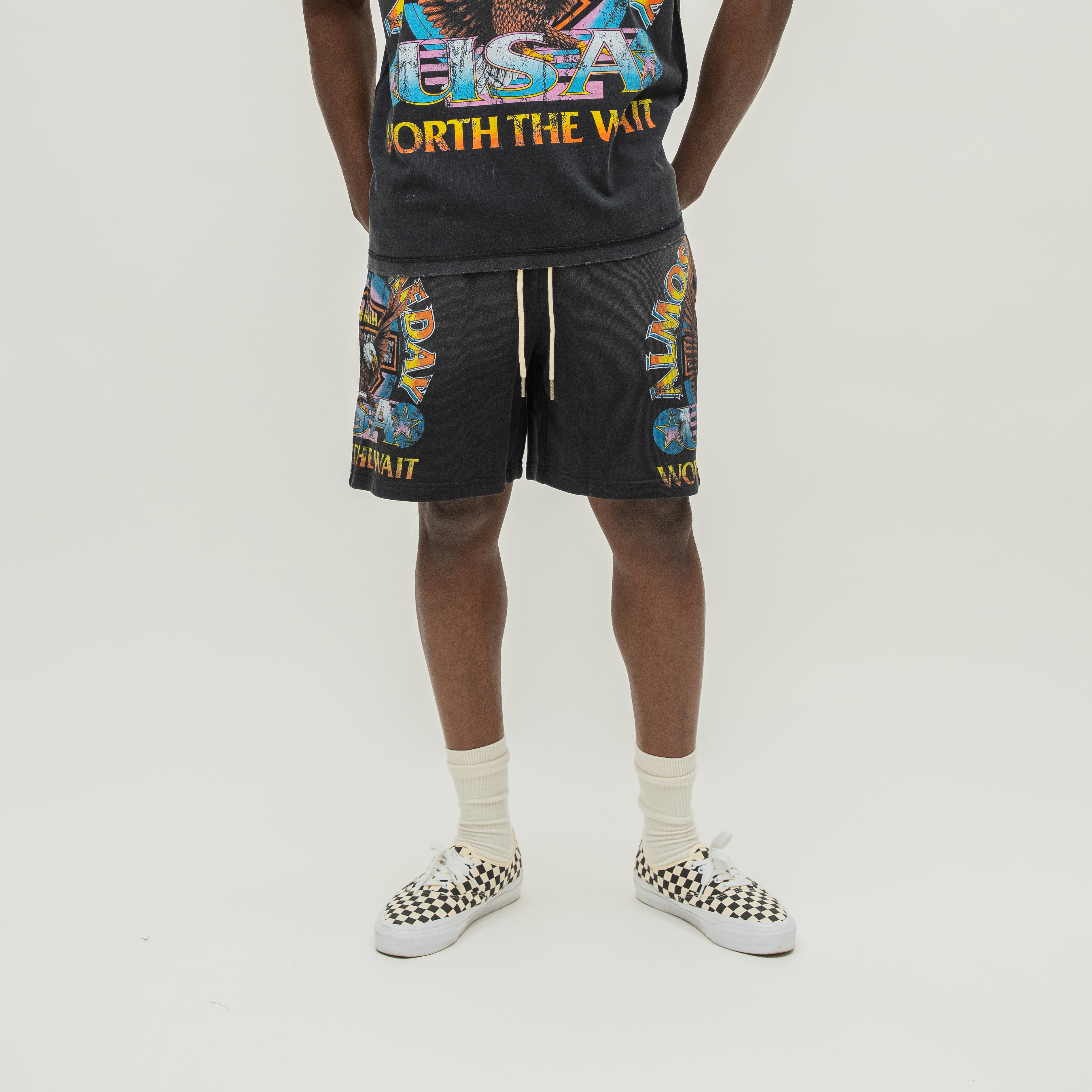 A model is shown from the waist down wearing black graphic shorts with colorful 'Worth the Wait' design, paired with a matching black graphic t-shirt, cream socks, and black-and-white checkered sneakers.