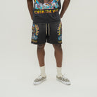 A model is shown from the waist down wearing black graphic shorts with colorful 'Worth the Wait' design, paired with a matching black graphic t-shirt, cream socks, and black-and-white checkered sneakers.