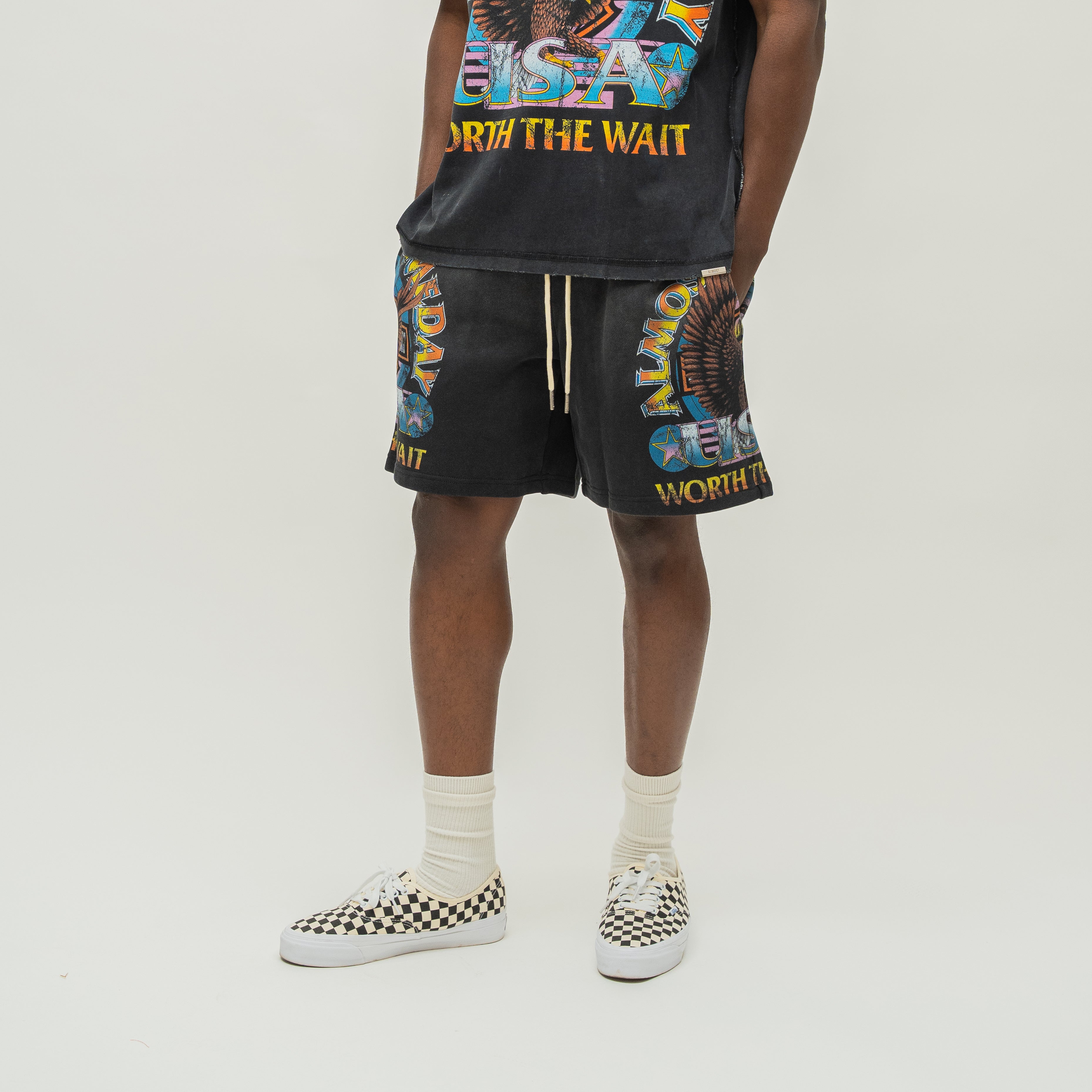 A model wearing a black graphic tank top and matching black graphic shorts with white crew socks and black-and-white checkered sneakers, photographed against a plain light gray studio background.