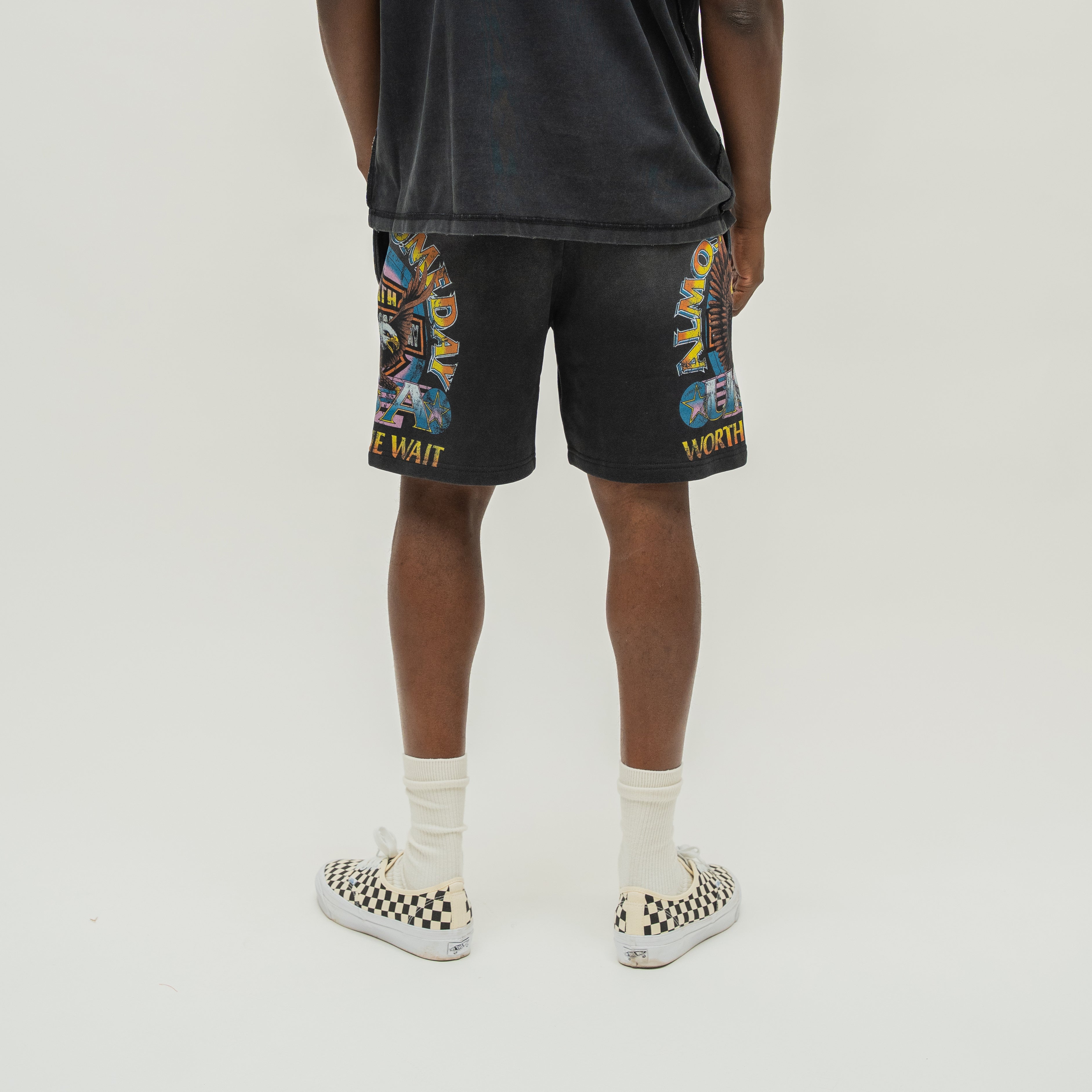 A person shown from the back wearing black graphic shorts with colorful print and yellow text, a faded black T-shirt, white socks, and black-and-white slip-on shoes against a plain light gray studio background.