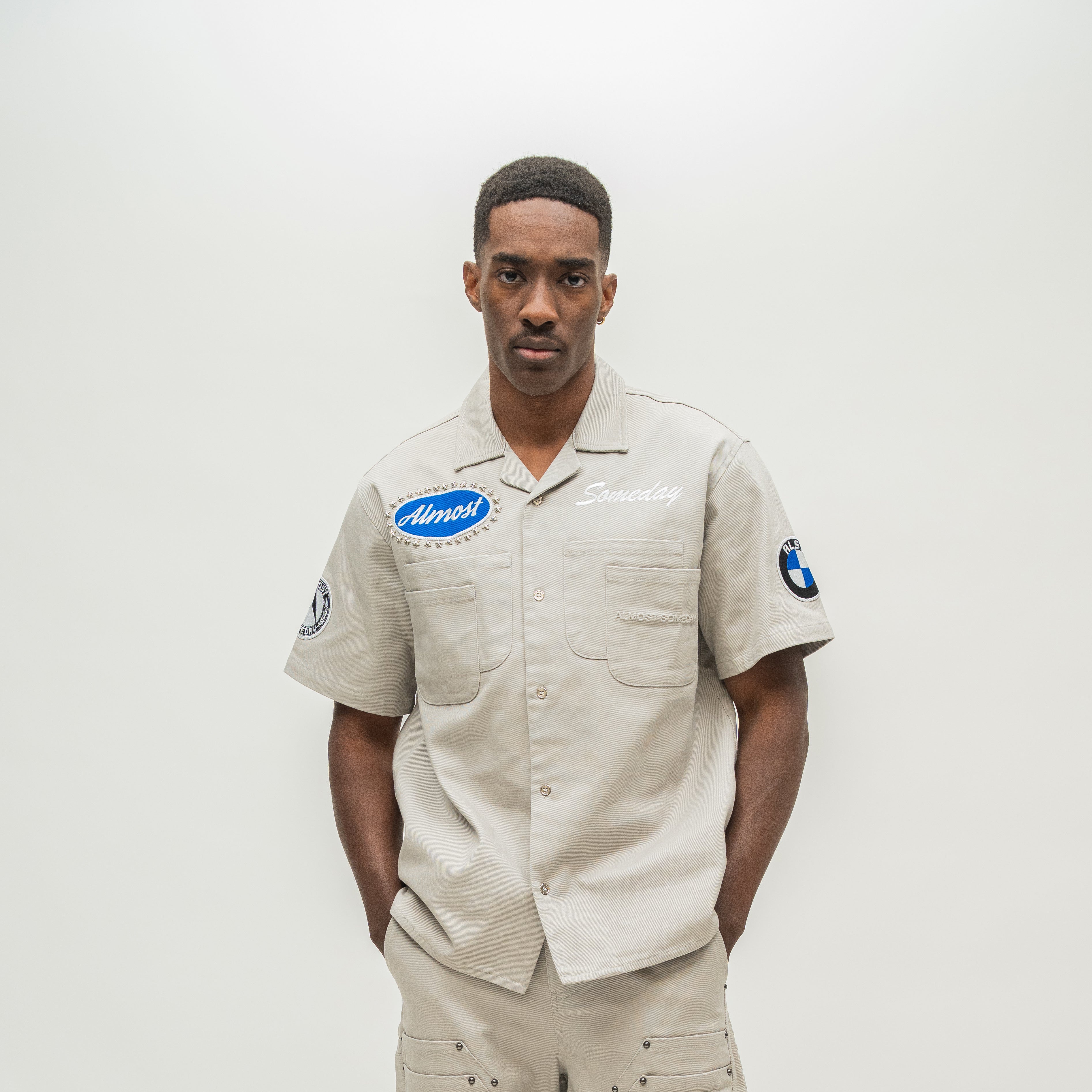 A man wearing a beige short-sleeve button-up work shirt with embroidered 'Almost' and 'Someday' logos on the chest, along with matching beige pants, standing against a plain light gray studio background.