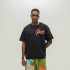 A man wearing a black oversized t-shirt with a red 'Almost Someday' logo on the chest, paired with olive green shorts featuring a tropical beach graphic and red lettering, set against a plain light gray background.
