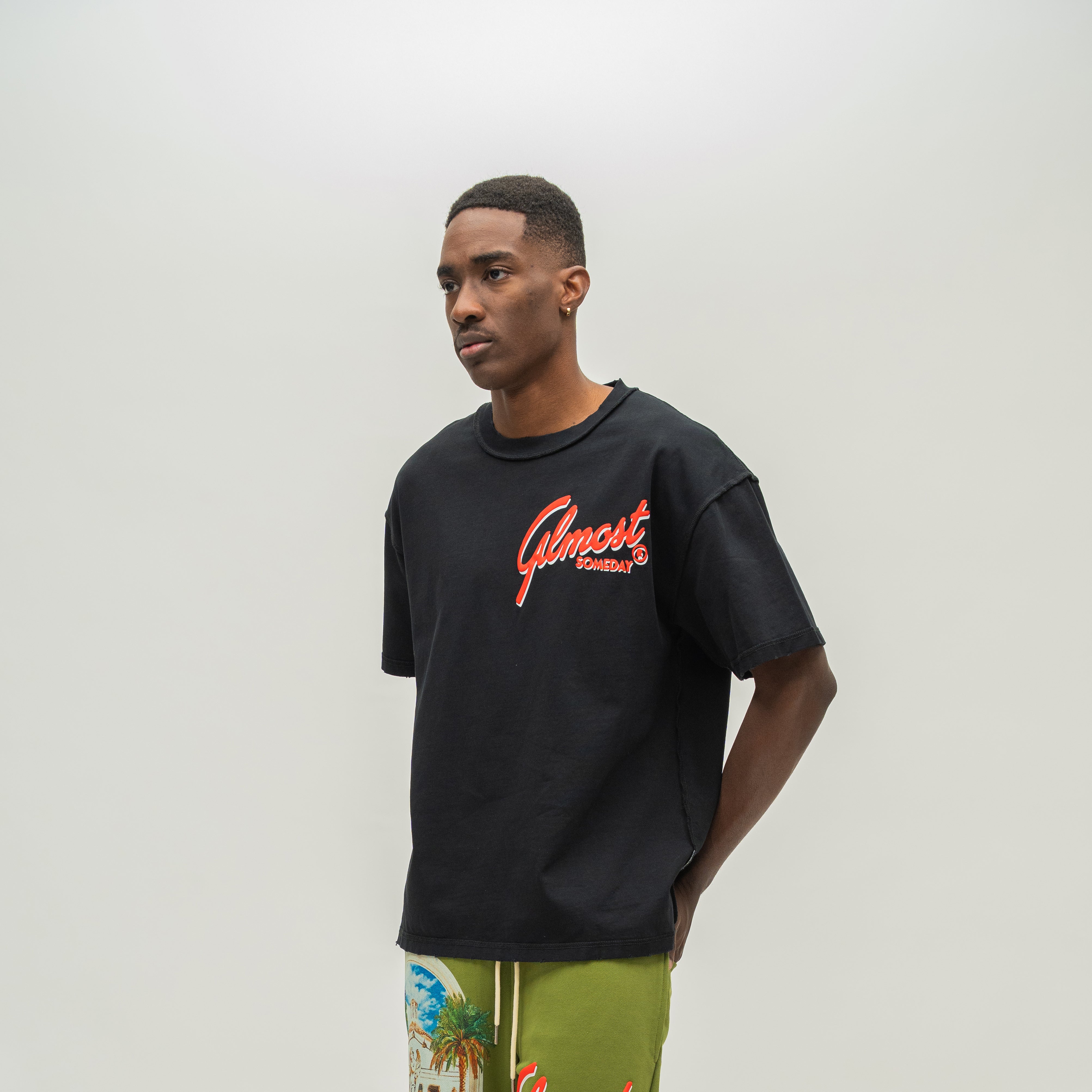 A man wearing a black crewneck t-shirt with a red and orange 'Almost Someday' graphic on the chest, paired with olive green tropical print shorts, standing against a plain light gray background.