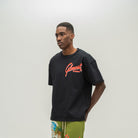 A man wearing a black crewneck t-shirt with a red and orange 'Almost Someday' graphic on the chest, paired with olive green tropical print shorts, standing against a plain light gray background.
