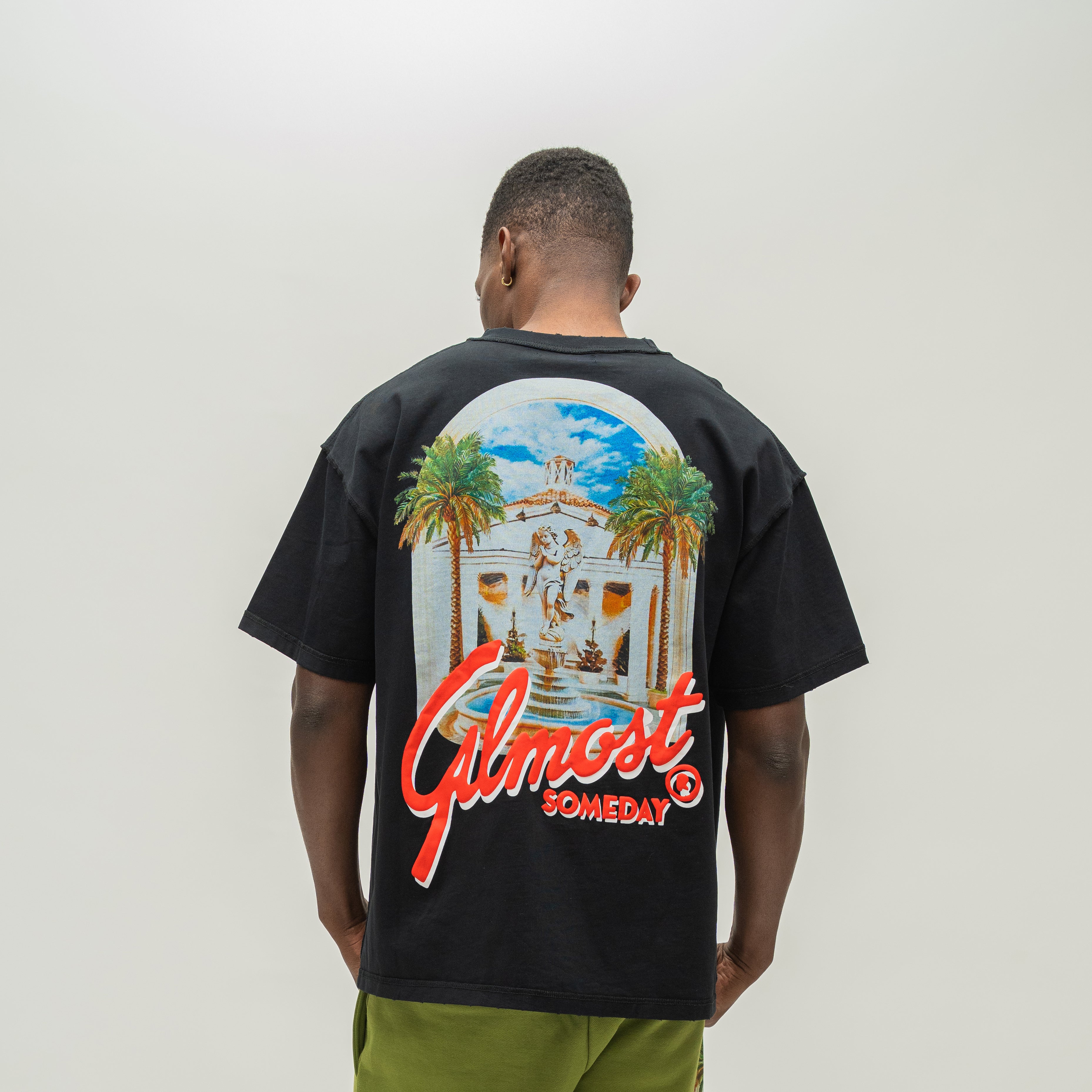 A man viewed from behind wearing a black t-shirt with a large graphic print on the back. The graphic features a white building with palm trees under a blue sky, with the red text 'ALMOST SOMEDAY' written across the bottom.
