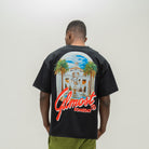 A man viewed from behind wearing a black t-shirt with a large graphic print on the back. The graphic features a white building with palm trees under a blue sky, with the red text 'ALMOST SOMEDAY' written across the bottom.