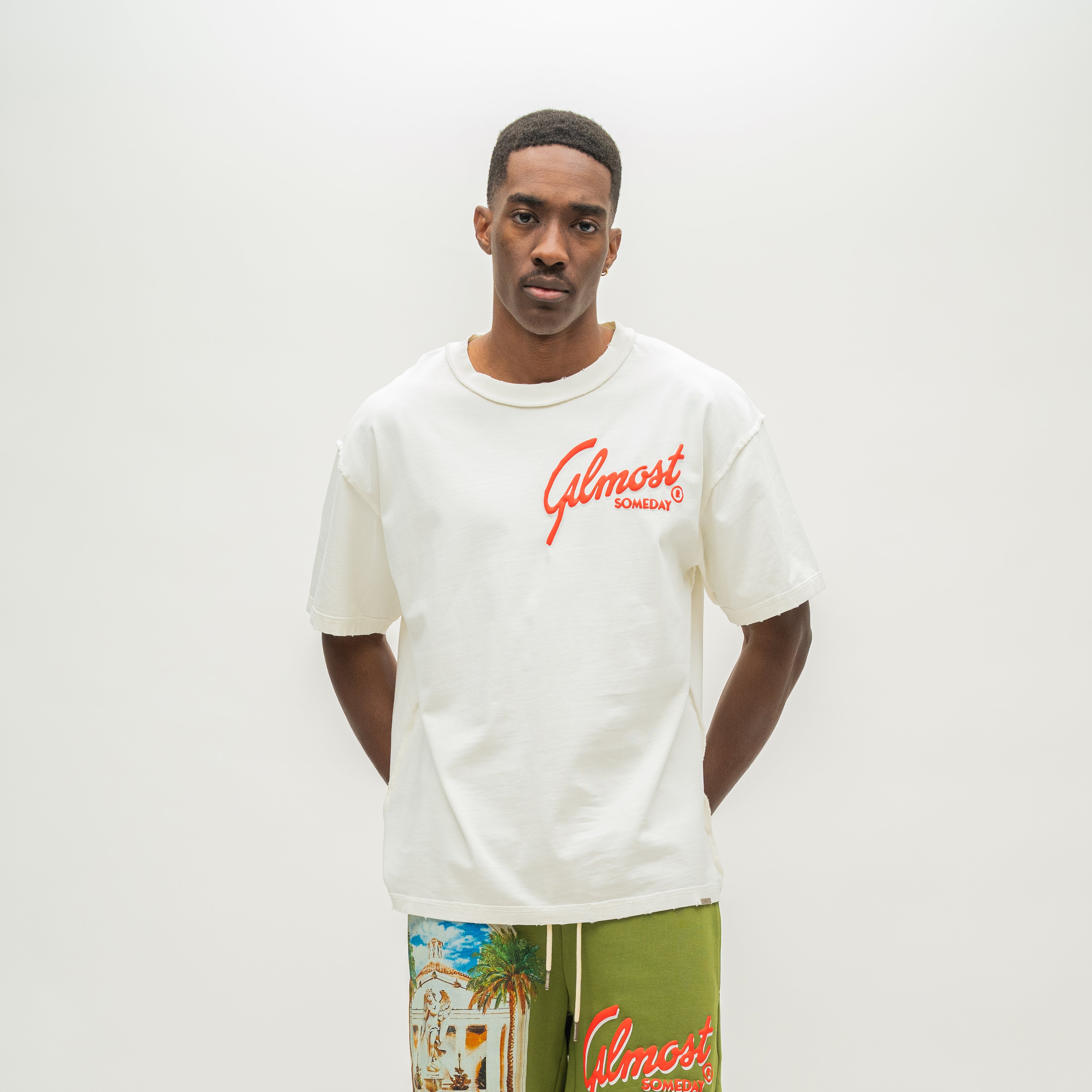 A man wearing a cream-colored t-shirt with red 'Almost Someday' script on the chest, paired with olive green shorts featuring a tropical beach print and the 'Almost Someday' logo. The background is a clean, off-white studio wall.