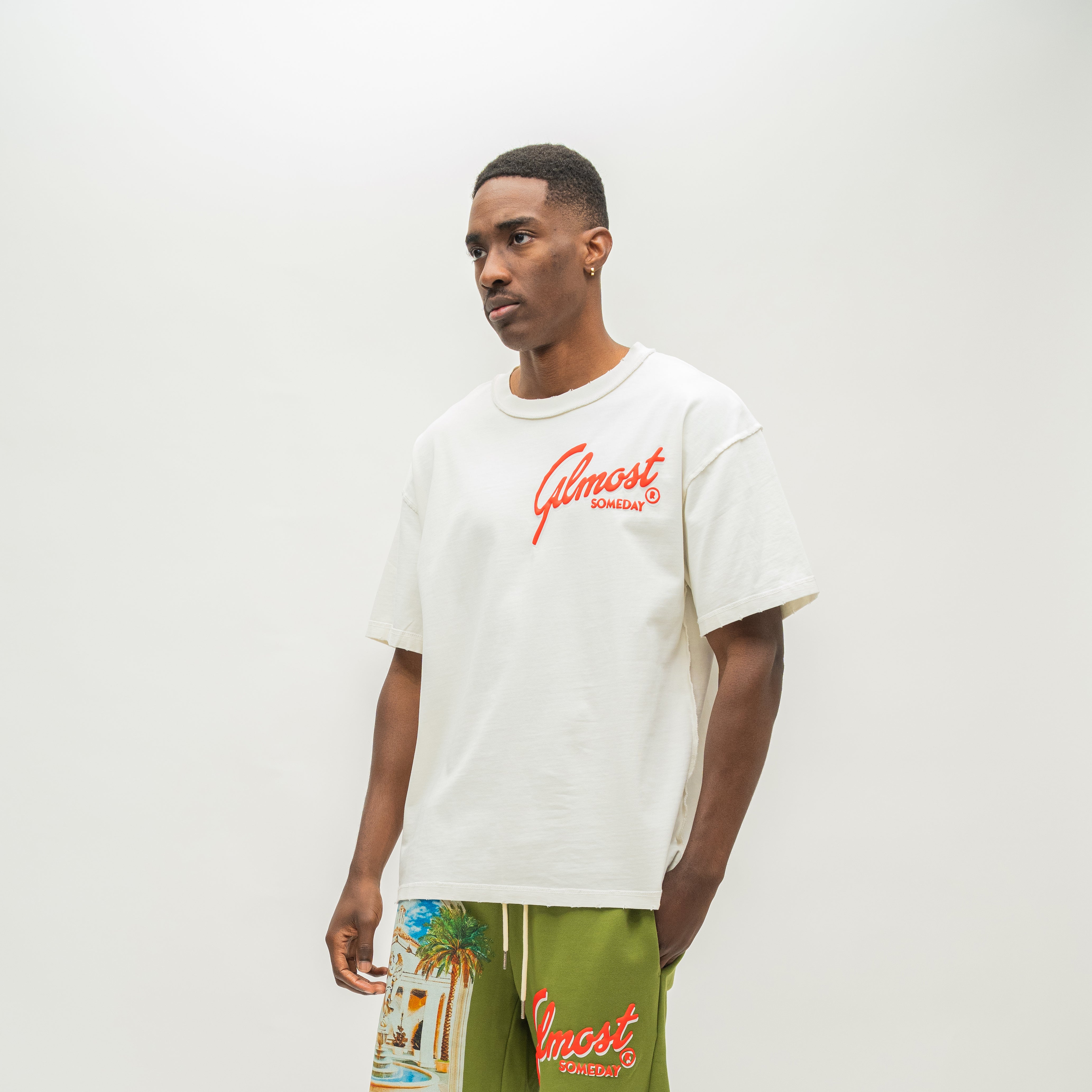 A man wearing an off-white t-shirt with a red 'Almost Someday' logo and green shorts with a beach graphic and red logo, standing against a plain light gray background.