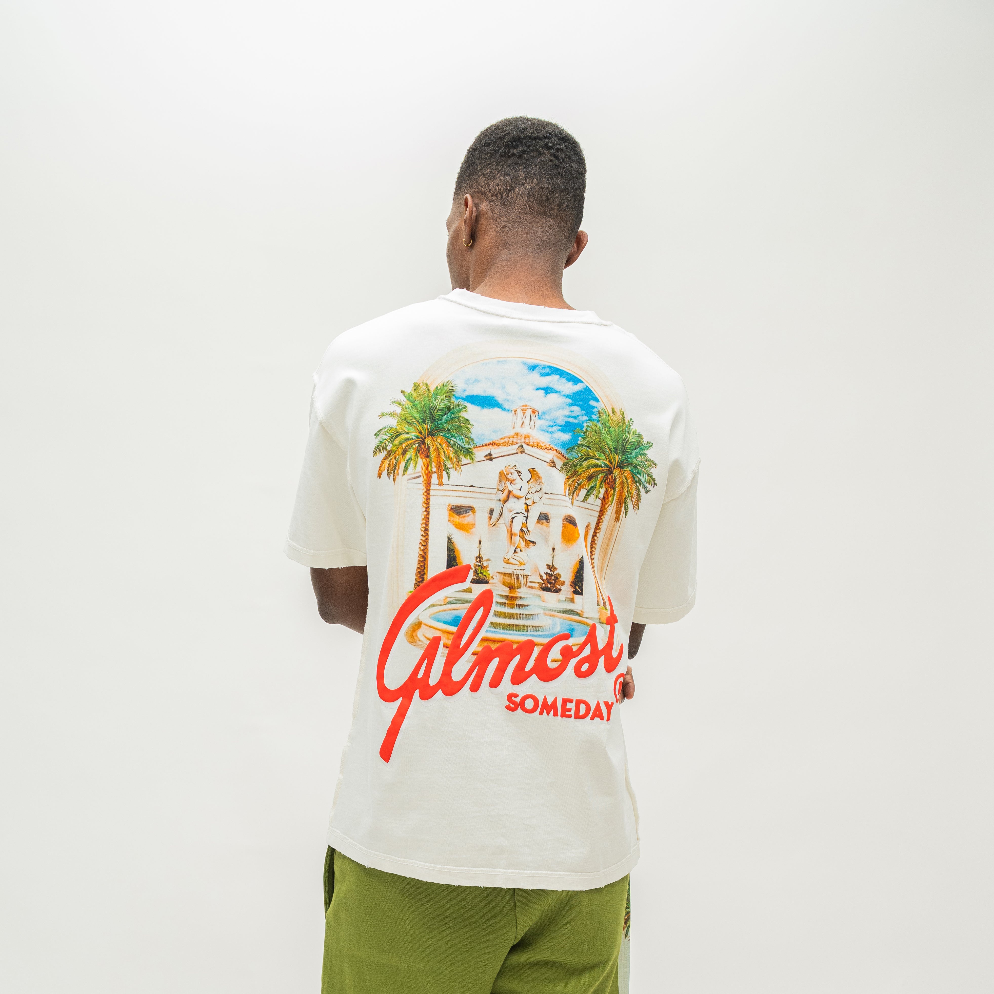 A man viewed from behind wearing a white t-shirt with a large colorful graphic on the back featuring palm trees, a building, and red script text reading 'Almost Someday', paired with olive green pants against a plain white background.