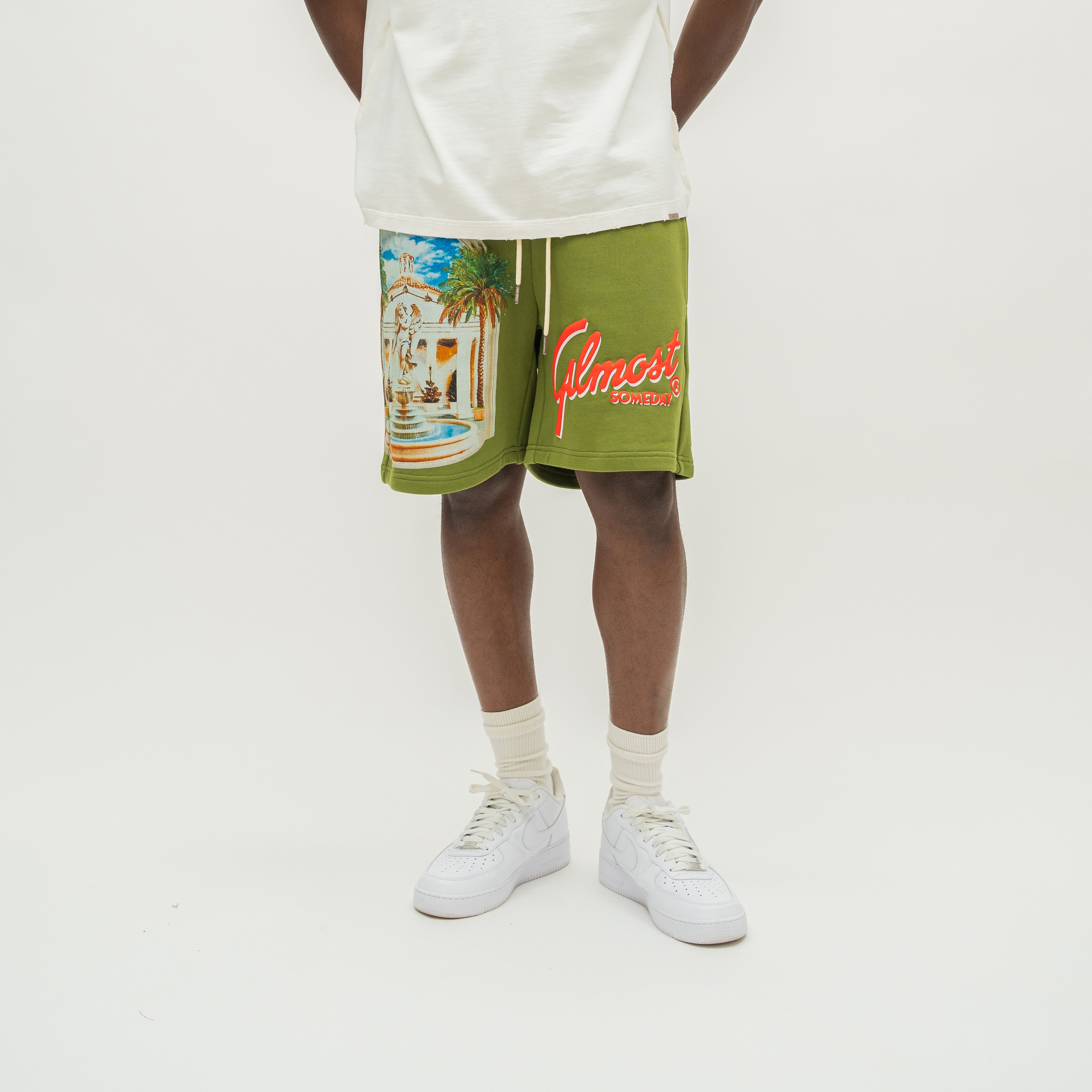 A person wearing olive green drawstring shorts with a large multicolor beach graphic on the left leg and red 'Almost Someday' script on the right leg, styled with a white tank top, white socks, and white sneakers against a plain white studio background.