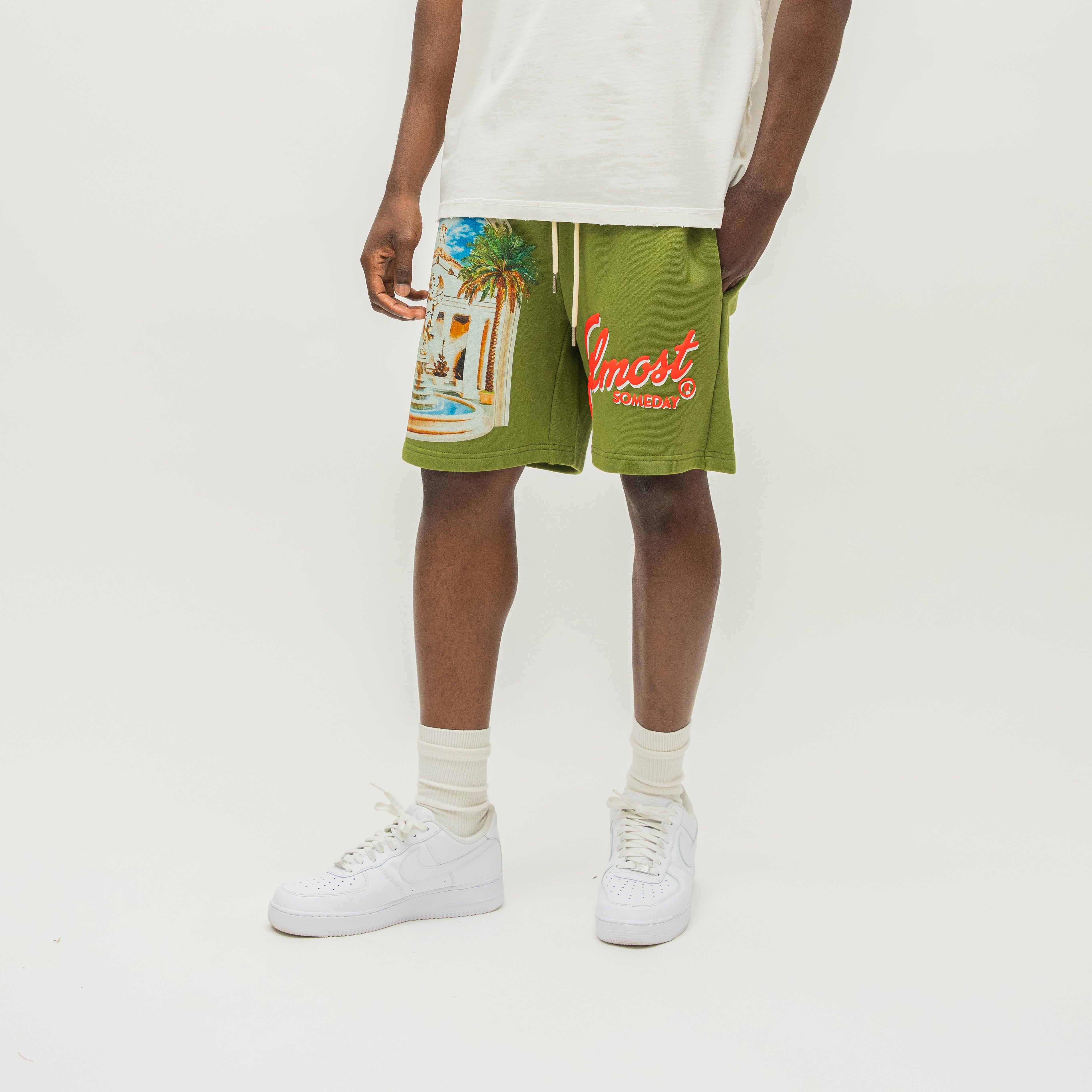 A person modeling olive green drawstring shorts with a colorful tropical beach graphic on the left leg and red 'Almost Someday' text on the right leg, paired with a white t-shirt, white socks, and white sneakers against a plain white studio background.